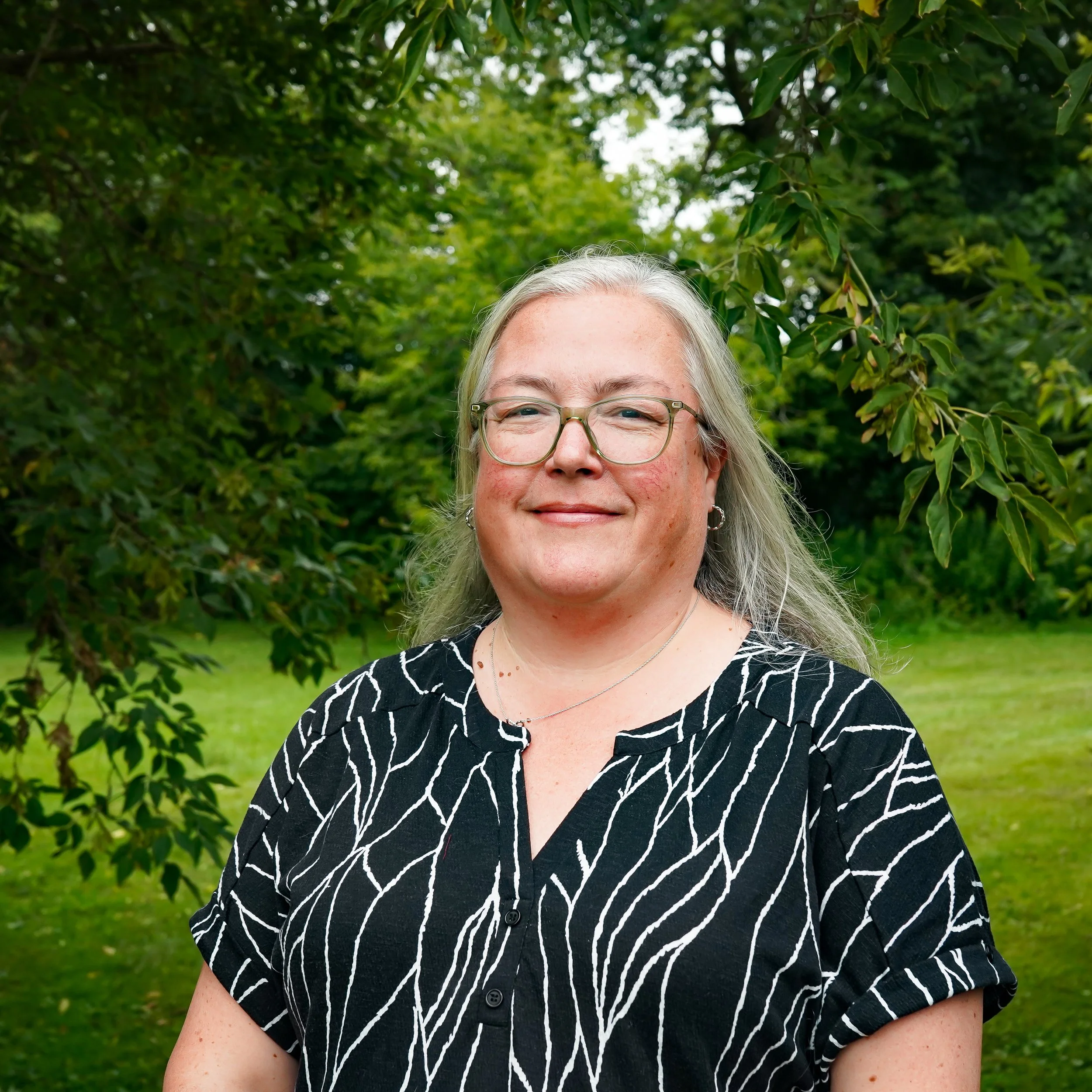 A woman with long gray hair, glasses, and a maroon top, smiling in front of a plain white background.