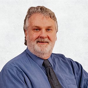 A middle-aged man with gray hair and a beard, wearing a blue shirt and dark tie, smiling at the camera.