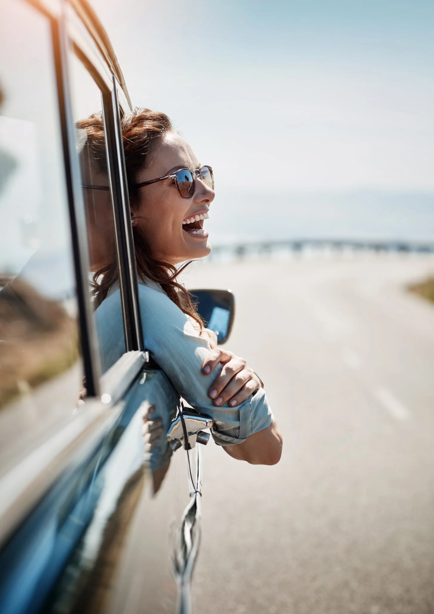 Woman with brown hair and sunglasses leaning out of car window, smiling, on a sunny day at the beach.