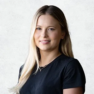 Close-up portrait of a young woman with long blonde hair, wearing a black shirt and smiling softly, standing against a light textured background.