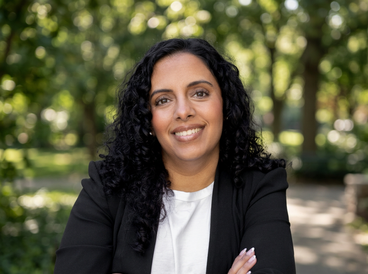 A confident woman with curly black hair wearing a black blazer and white top, smiling with arms crossed, standing against a light background.