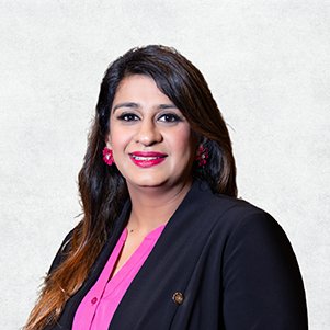 Headshot of a woman with medium-length dark hair, wearing pink lipstick, pink earrings, a pink blouse, and a black blazer against a light gray background.