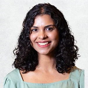 A woman with shoulder-length curly dark hair, smiling, wearing a light green blouse, standing against a plain white background.