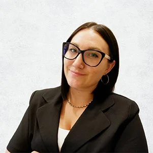 A young woman with short dark hair, glasses, a nose ring, and hoop earrings, smiling and posing for the camera against a plain light background, wearing a black blazer and white top.
