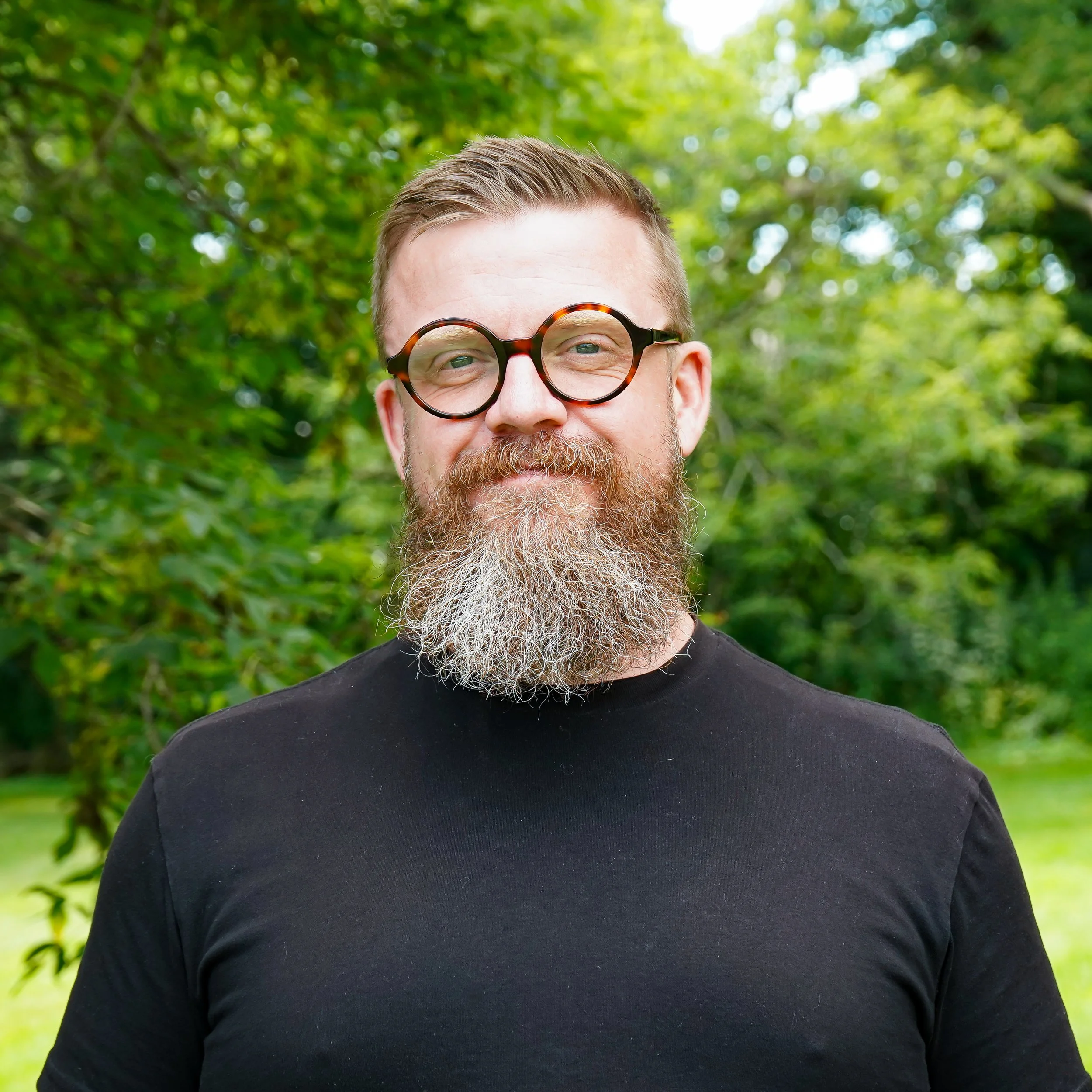 A man with a beard and short hair standing outdoors against a gray background, wearing a black t-shirt.