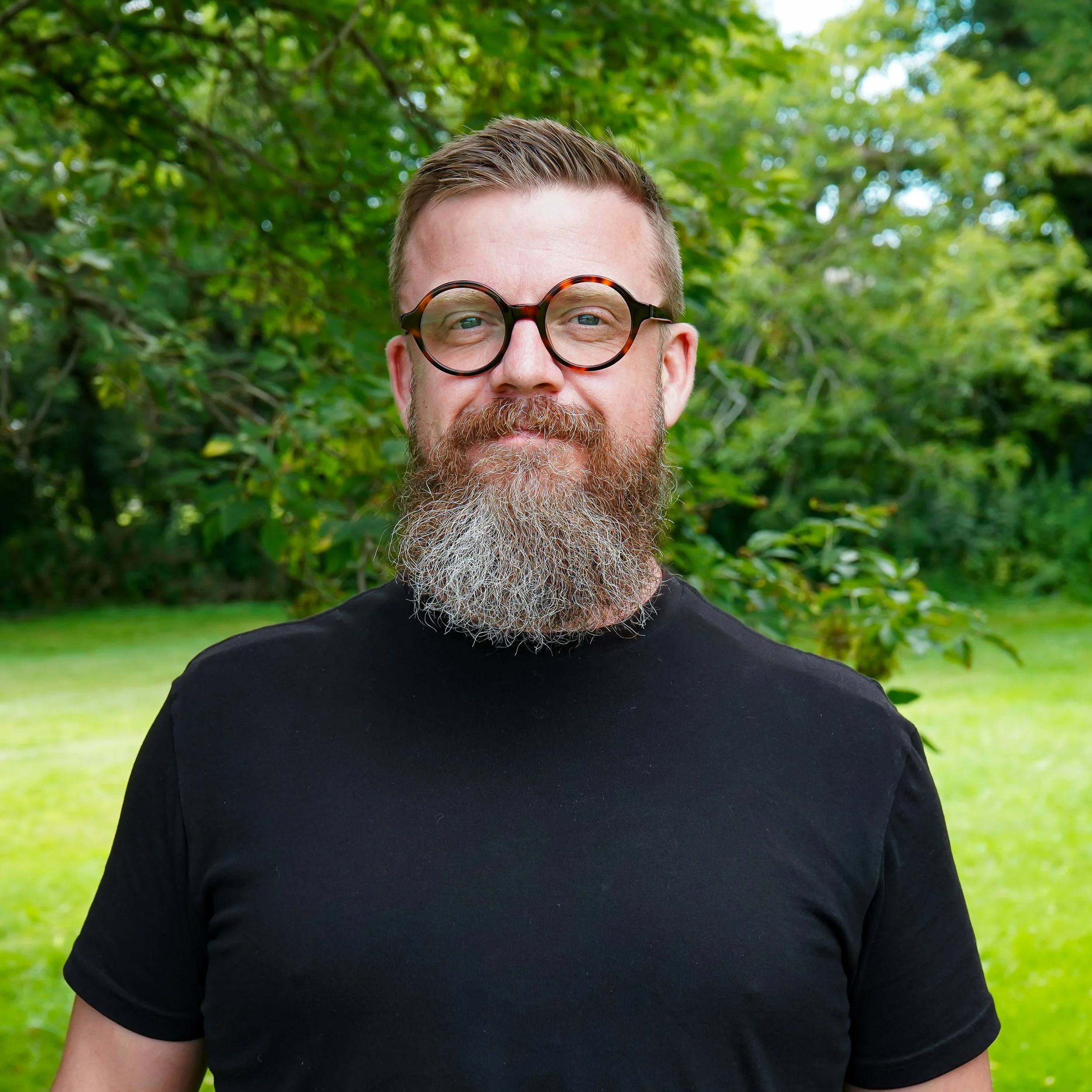A man with a beard and short hair standing outdoors against a gray background, wearing a black t-shirt.