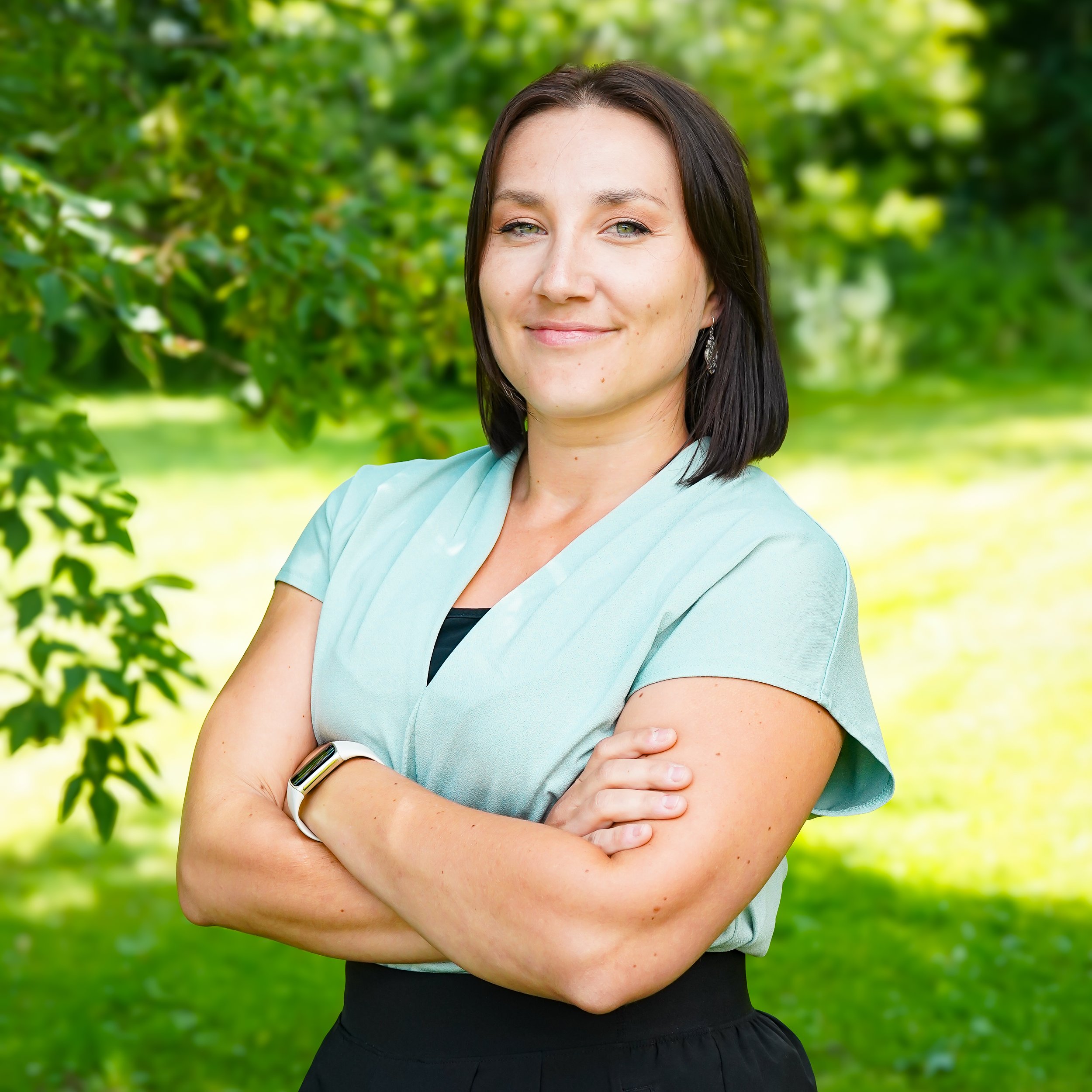 A woman with dark hair, wearing earrings, a light green shirt, and dark pants, standing outdoors with her arms crossed in front of greenery.