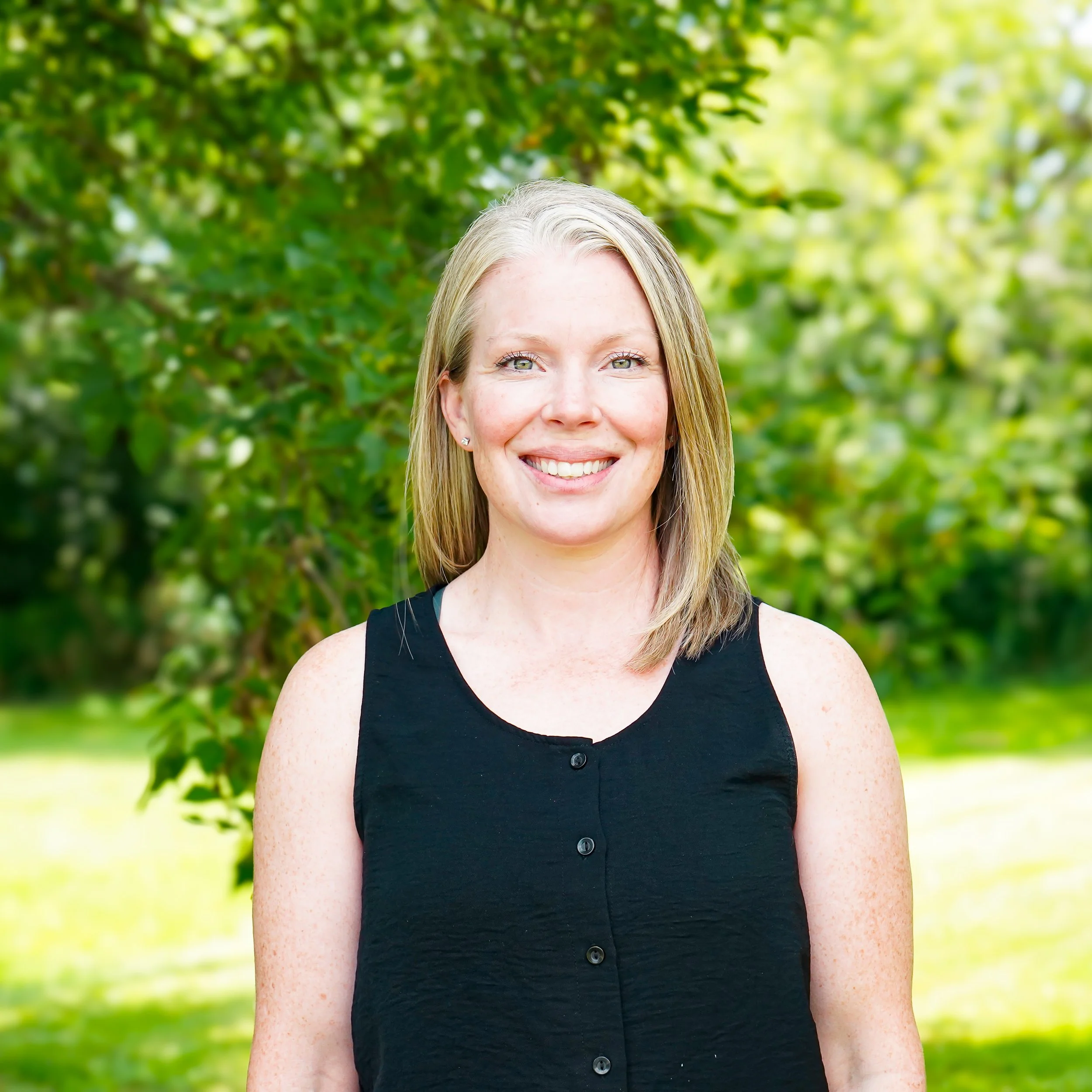 A woman with straight blonde hair, blue eyes, and a black sleeveless top smiling at the camera against a plain white background.