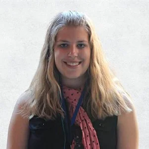 A young woman with long, wavy blonde hair smiling at the camera, wearing a black top and a pink scarf with black polka dots, against a plain white background.