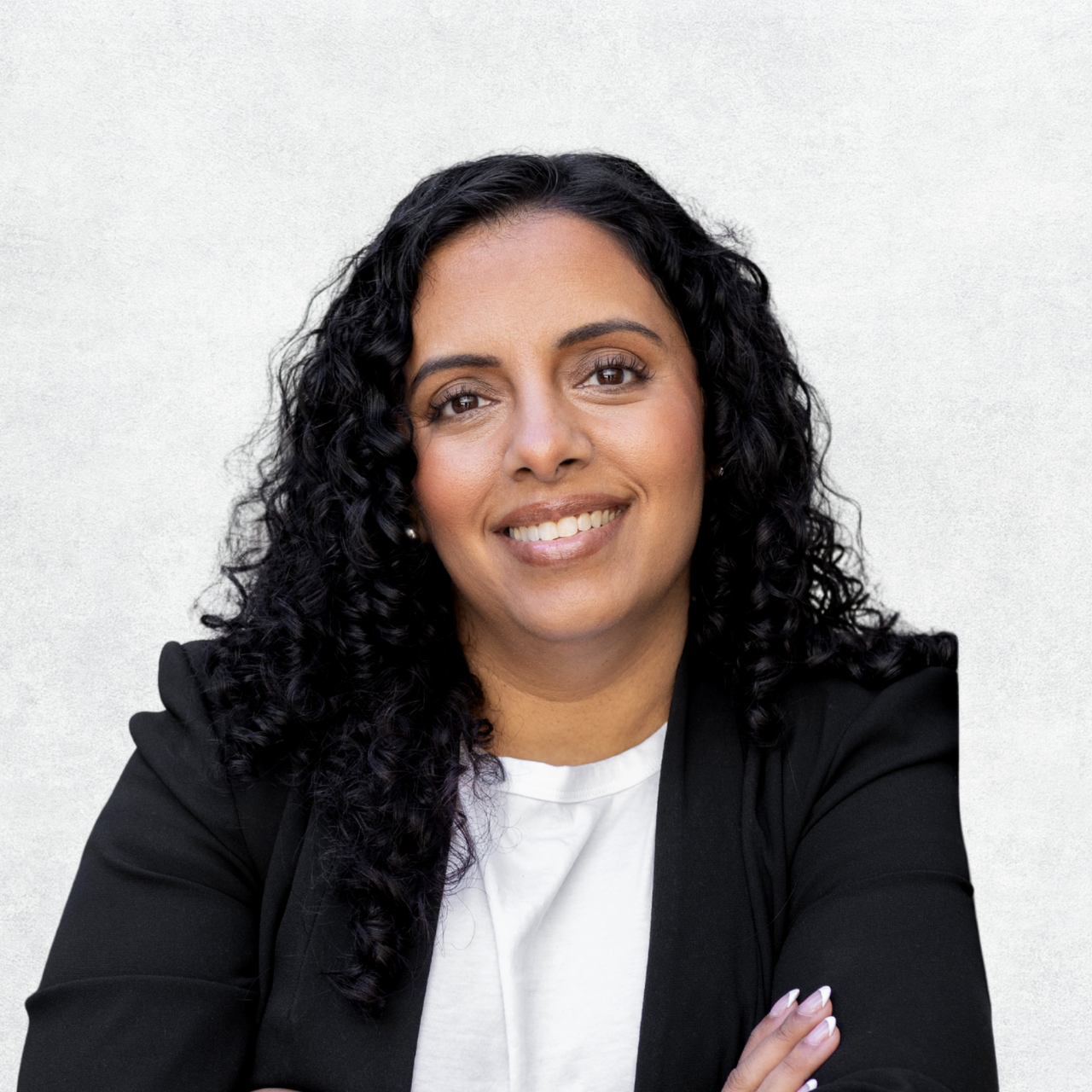 A confident woman with curly black hair wearing a black blazer and white top, smiling with arms crossed, standing against a light background.