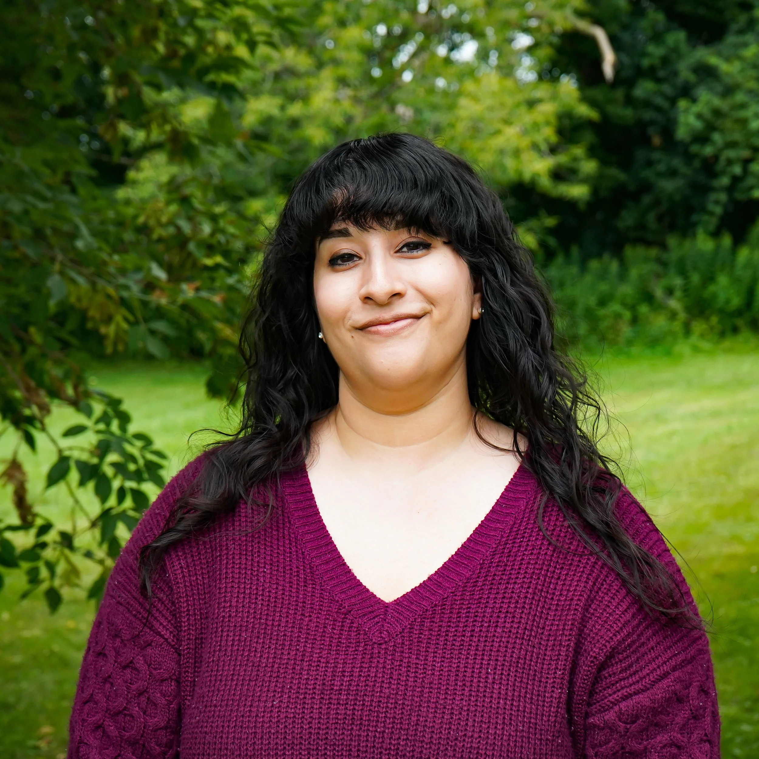 Close-up of a smiling woman with dark, curly hair wearing a purple knit sweater, standing outdoors with green foliage in the background.