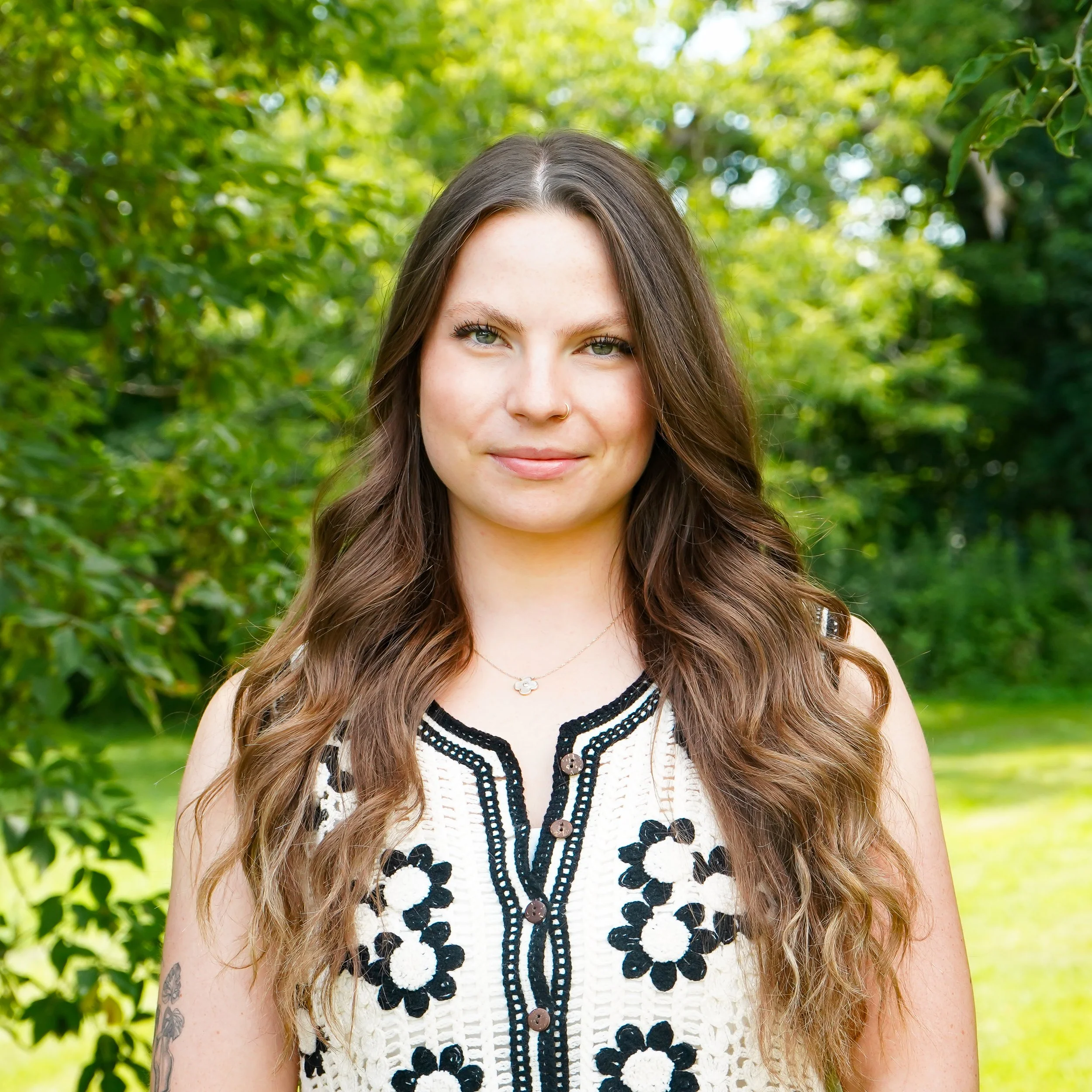 Close-up of a woman with long, wavy brown hair and freckles, wearing a white crochet top with black floral embroidery, standing outdoors with green foliage background.