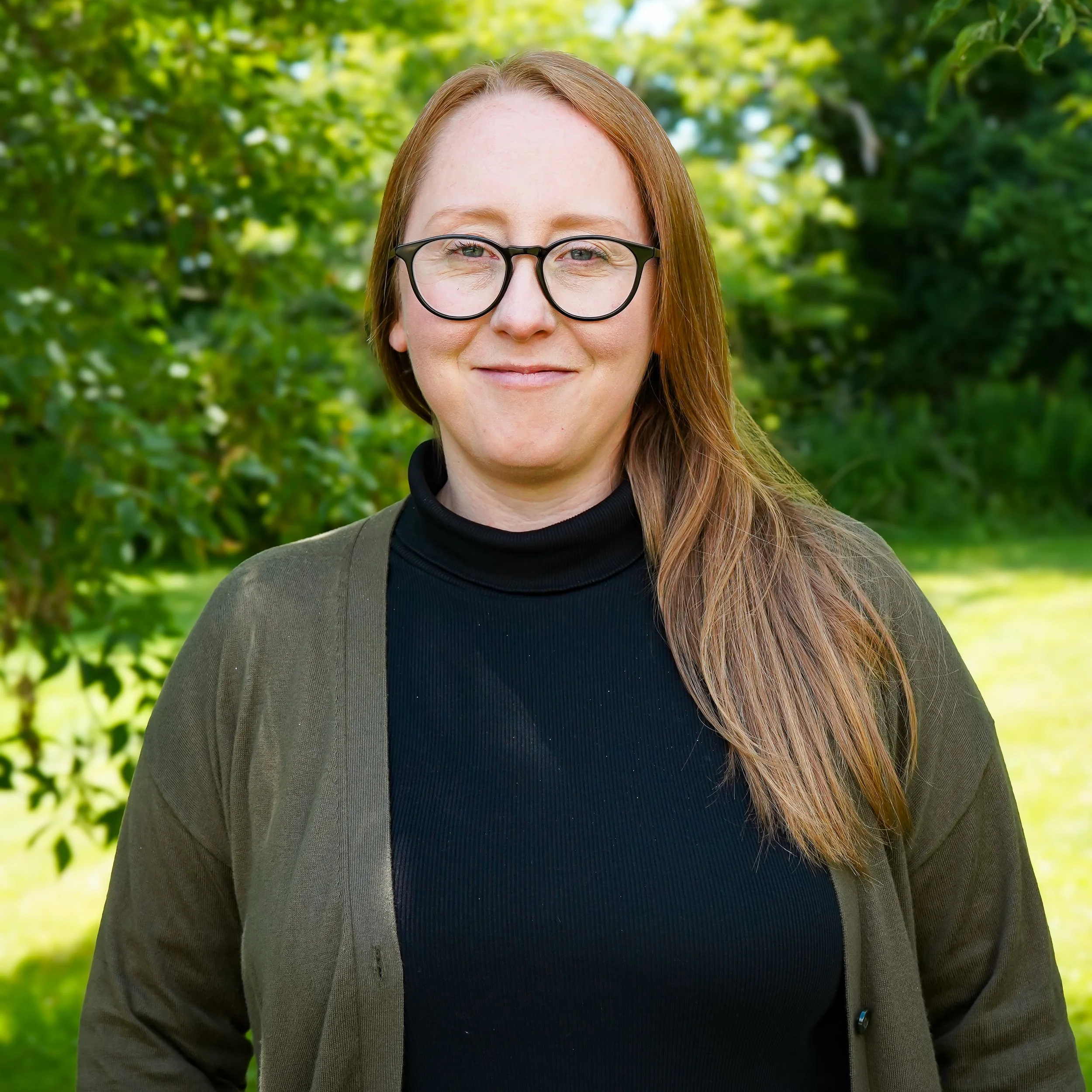 A woman with long red hair, wearing glasses and a navy blue sweater, smiling at the camera against a light textured background.