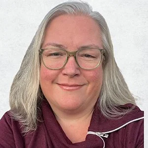 A woman with long gray hair, glasses, and a maroon top, smiling in front of a plain white background.
