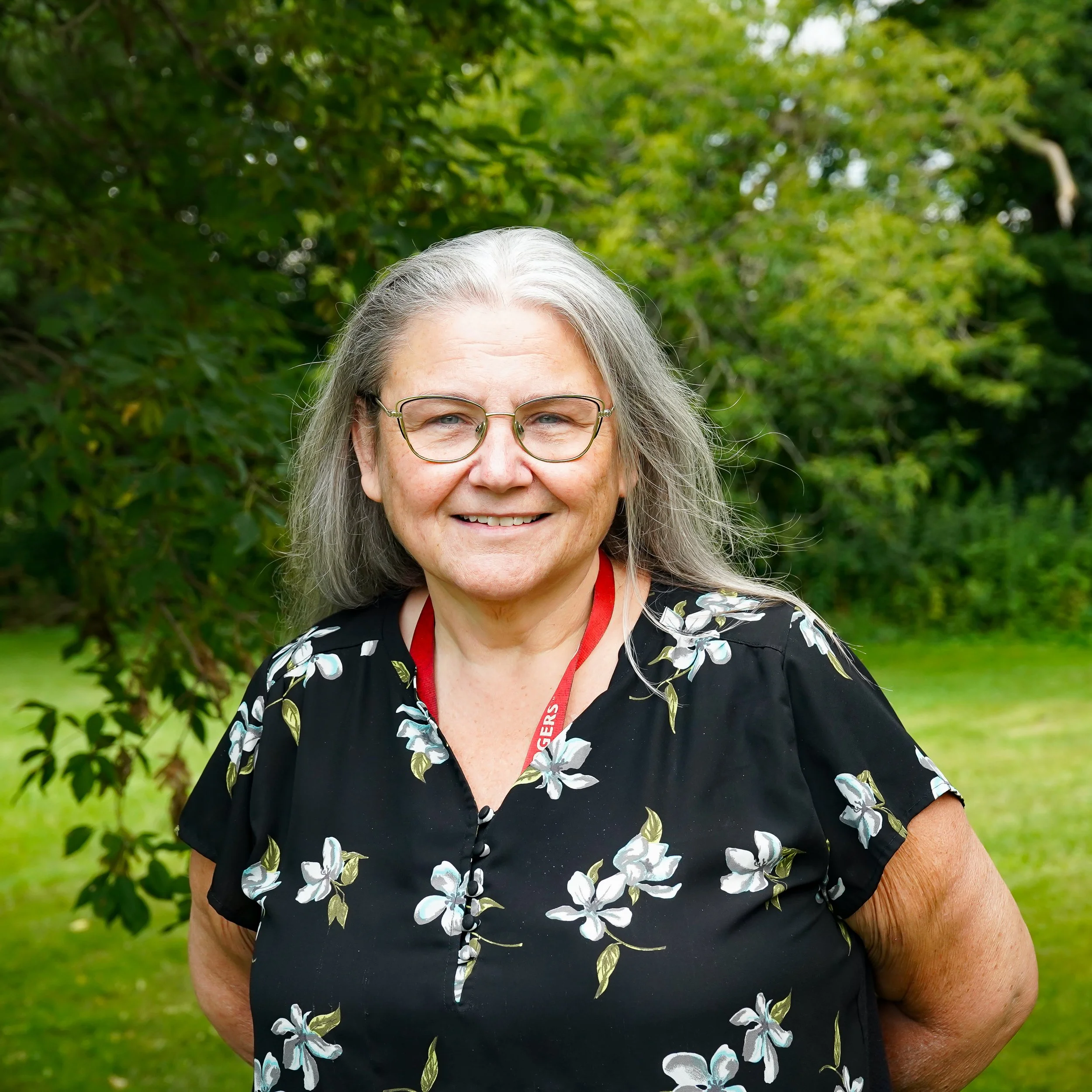 Close-up of an older woman with gray hair and glasses smiling outdoors with green trees in the background.