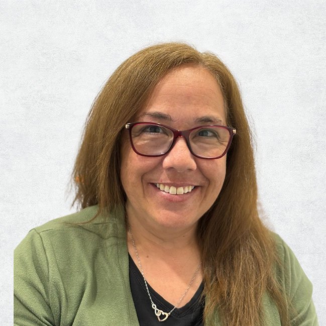 A smiling woman with long brown hair, wearing glasses, a green jacket, and a black top, smiling against a light background.