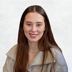 A young woman with long brown hair smiling at the camera, wearing a beige jacket and gold earrings, against a plain light background.
