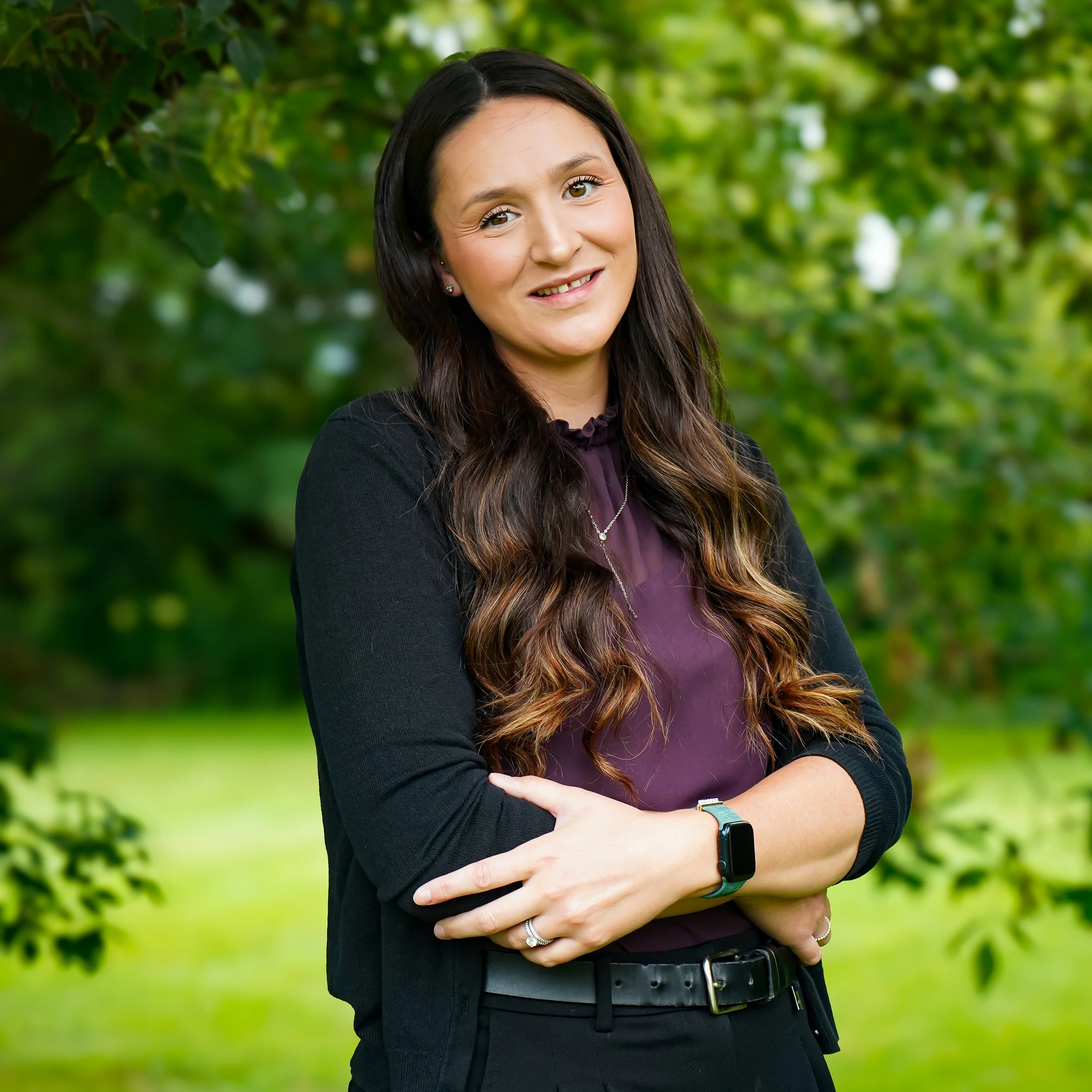 A woman with long brown hair, wearing a black blazer and purple blouse, standing outdoors with her arms crossed, in front of green trees.