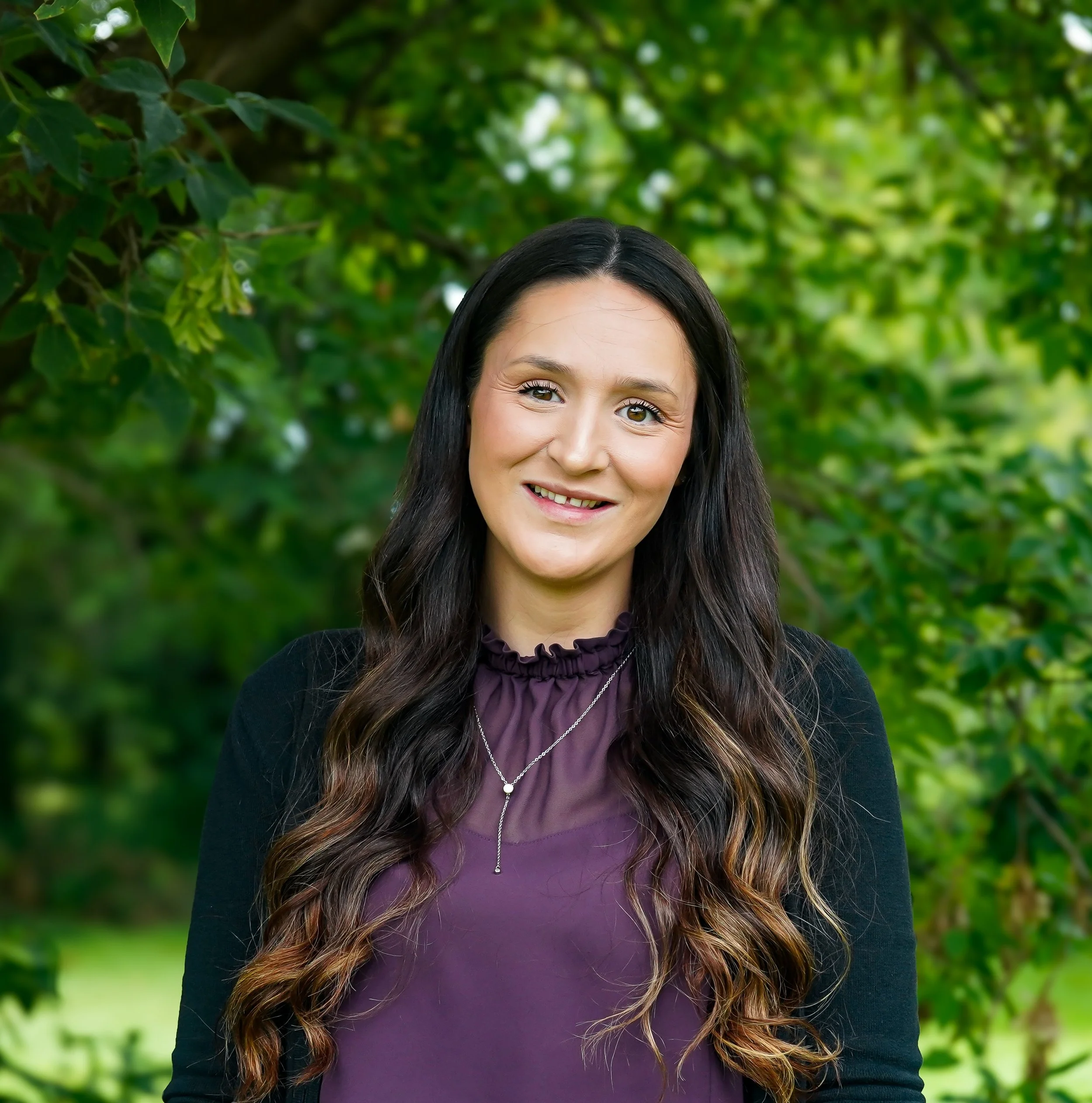 Portrait of a young woman with long wavy brown hair, wearing a black top and purple cardigan, smiling softly at the camera against a plain white background.