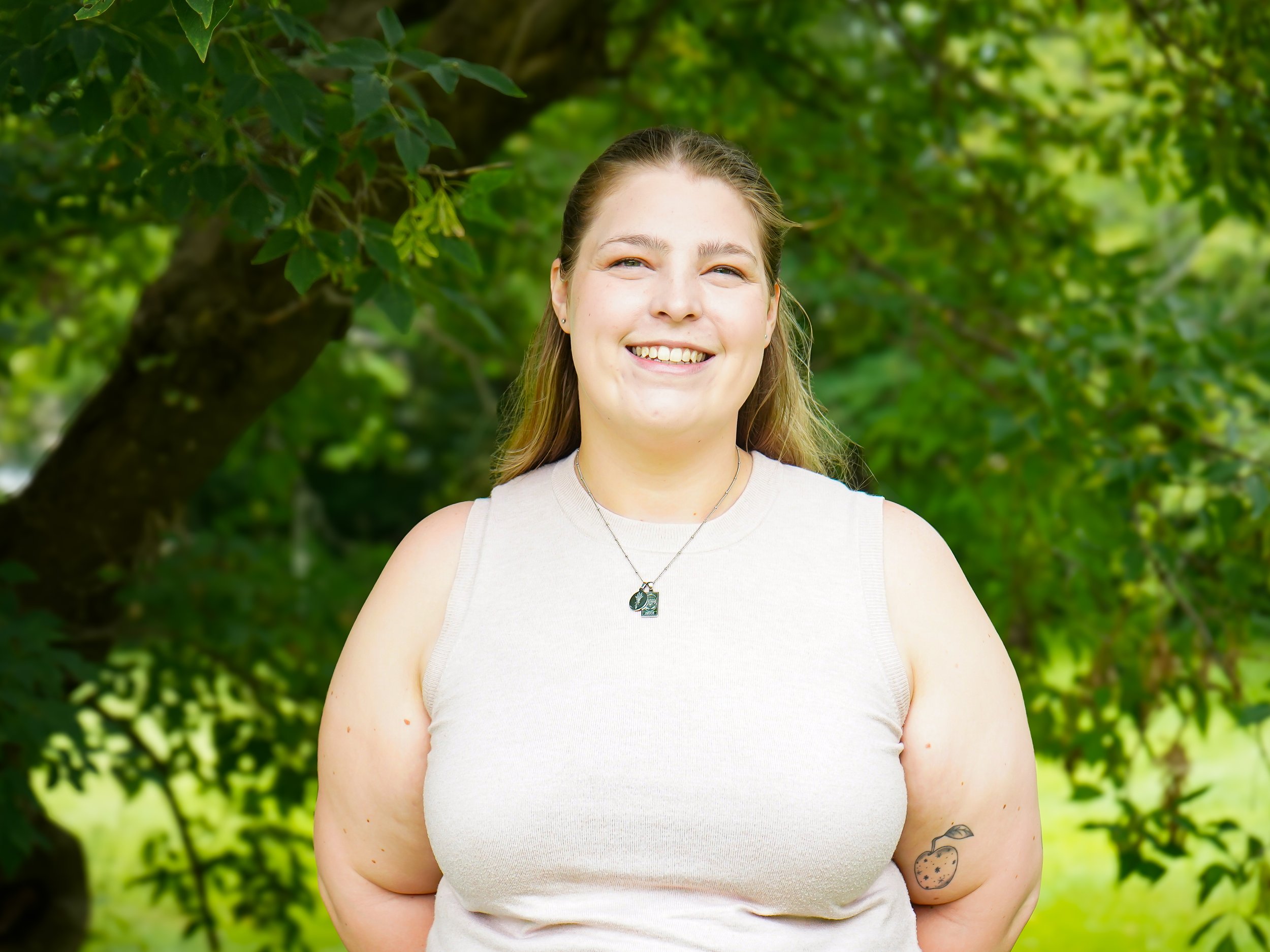 A young woman with long, light brown hair and a tattoo of a strawberry on her left arm, smiling outdoors in front of green foliage and a tree.