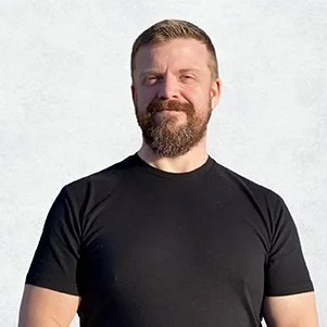 A man with a beard and short hair standing outdoors against a gray background, wearing a black t-shirt.