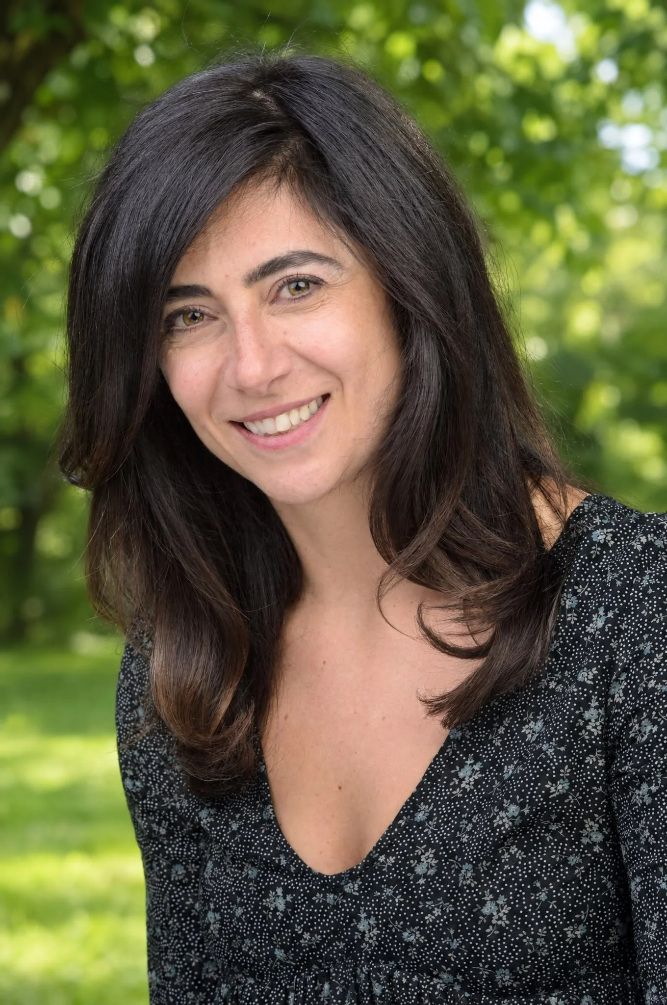 A woman with long dark hair, smiling, wearing a black patterned top, with a light gray background.