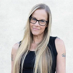 A woman with long blonde hair, wearing black glasses and a black sleeveless top, smiling slightly against a plain light background.