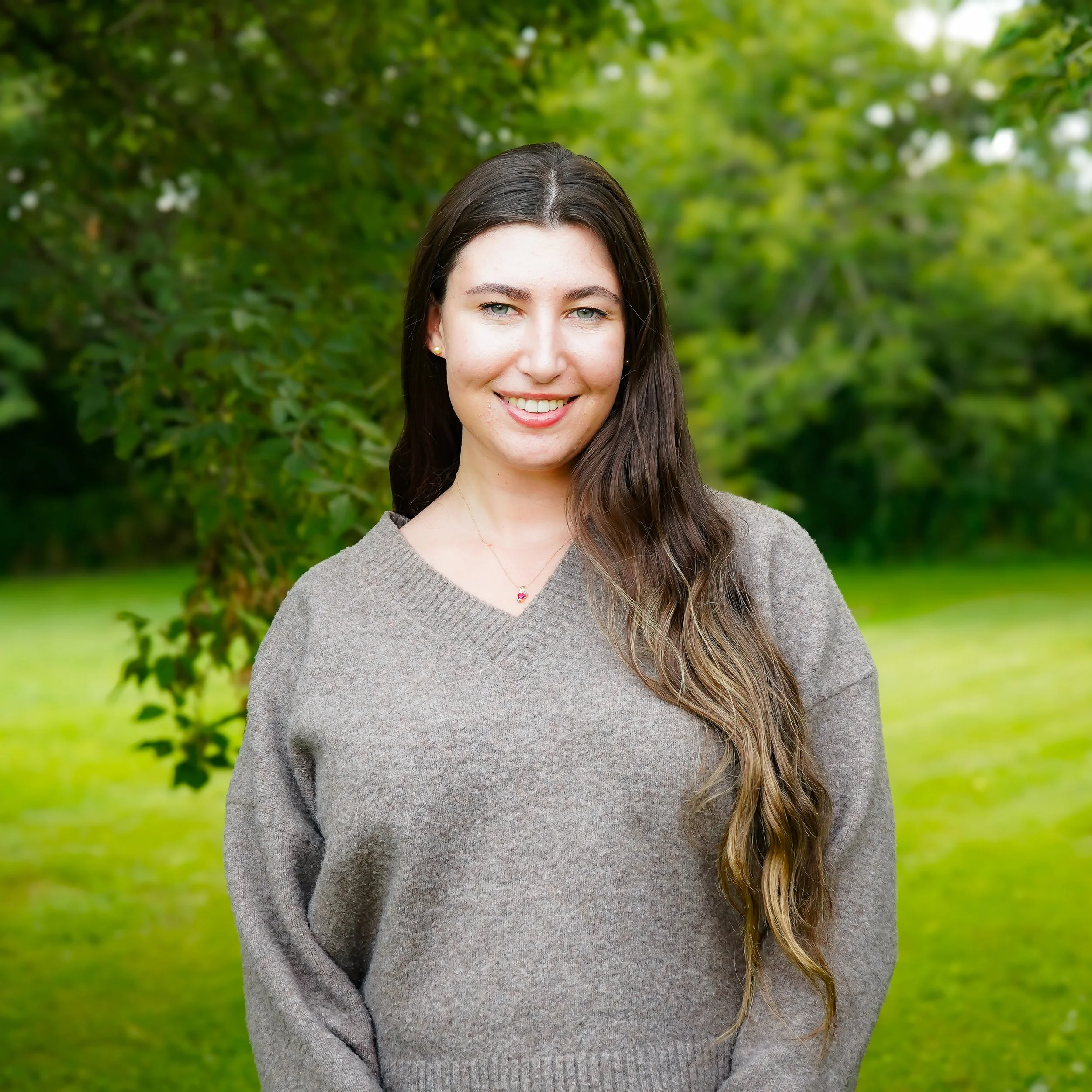 A young woman with long brown hair, smiling, wearing a gray sweater and a necklace, standing outdoors in front of green trees.