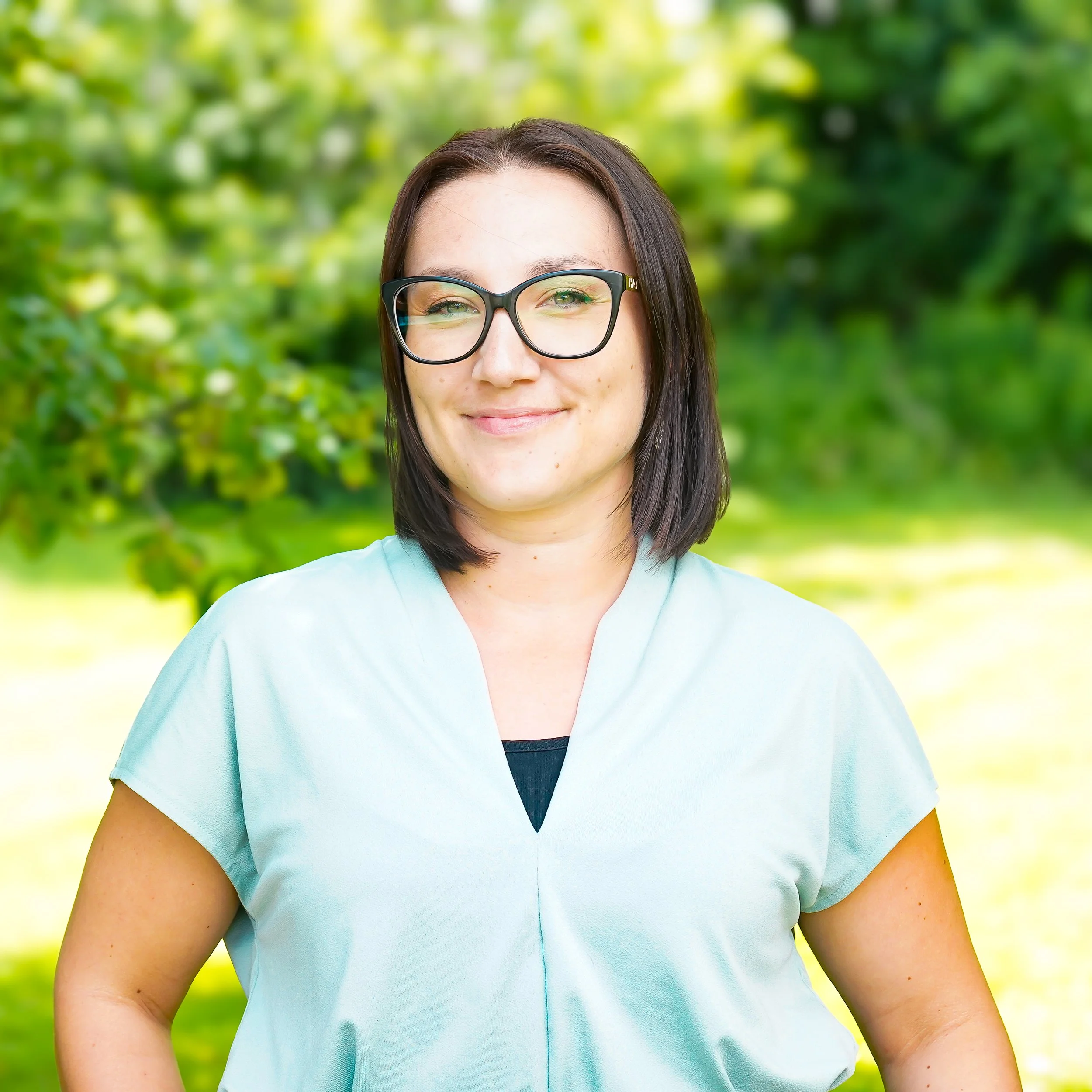 A young woman with short dark hair, glasses, a nose ring, and hoop earrings, smiling and posing for the camera against a plain light background, wearing a black blazer and white top.