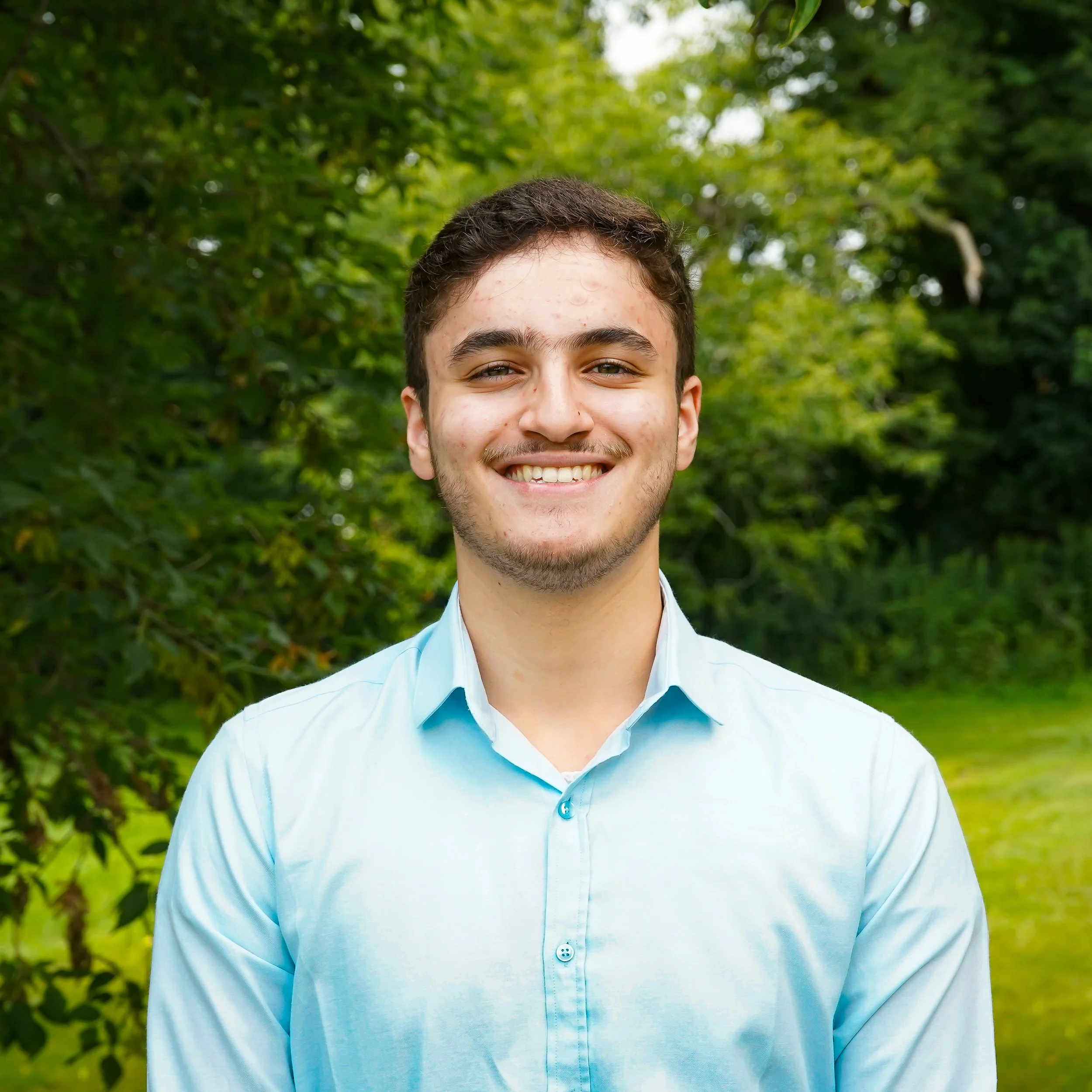 Close-up of a young man's smiling face with short dark hair, wearing a light blue shirt, outdoors with green foliage in the background.