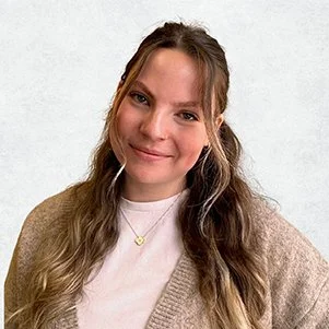A young woman with long, wavy brown hair and blue eyes, smiling, wearing a beige cardigan over a white top, with a gold necklace featuring a flower-shaped pendant, standing against a plain light gray background.