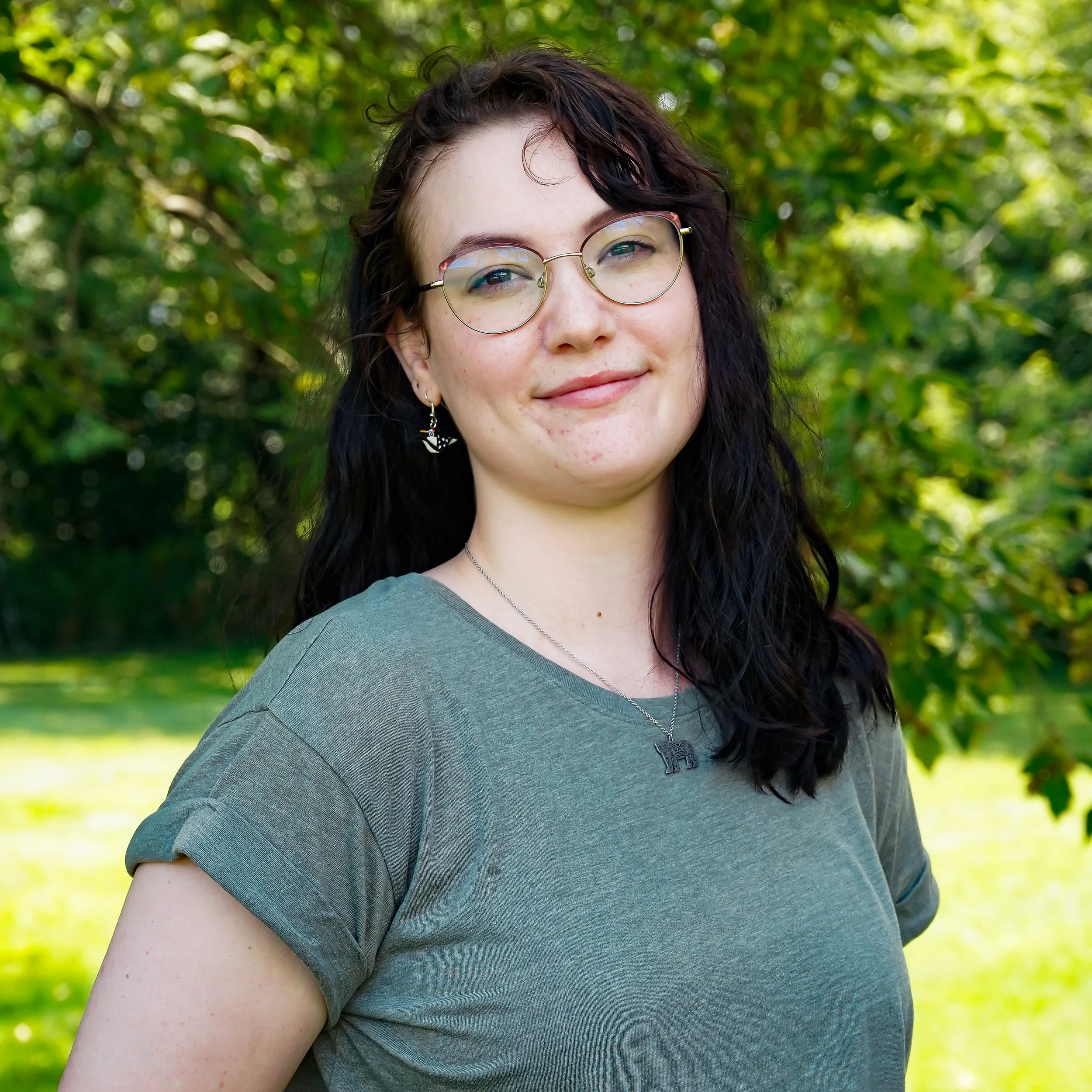 A young woman with long black wavy hair, wearing glasses, earrings, a necklace, and a brown knitted sweater, smiling against a plain white background.