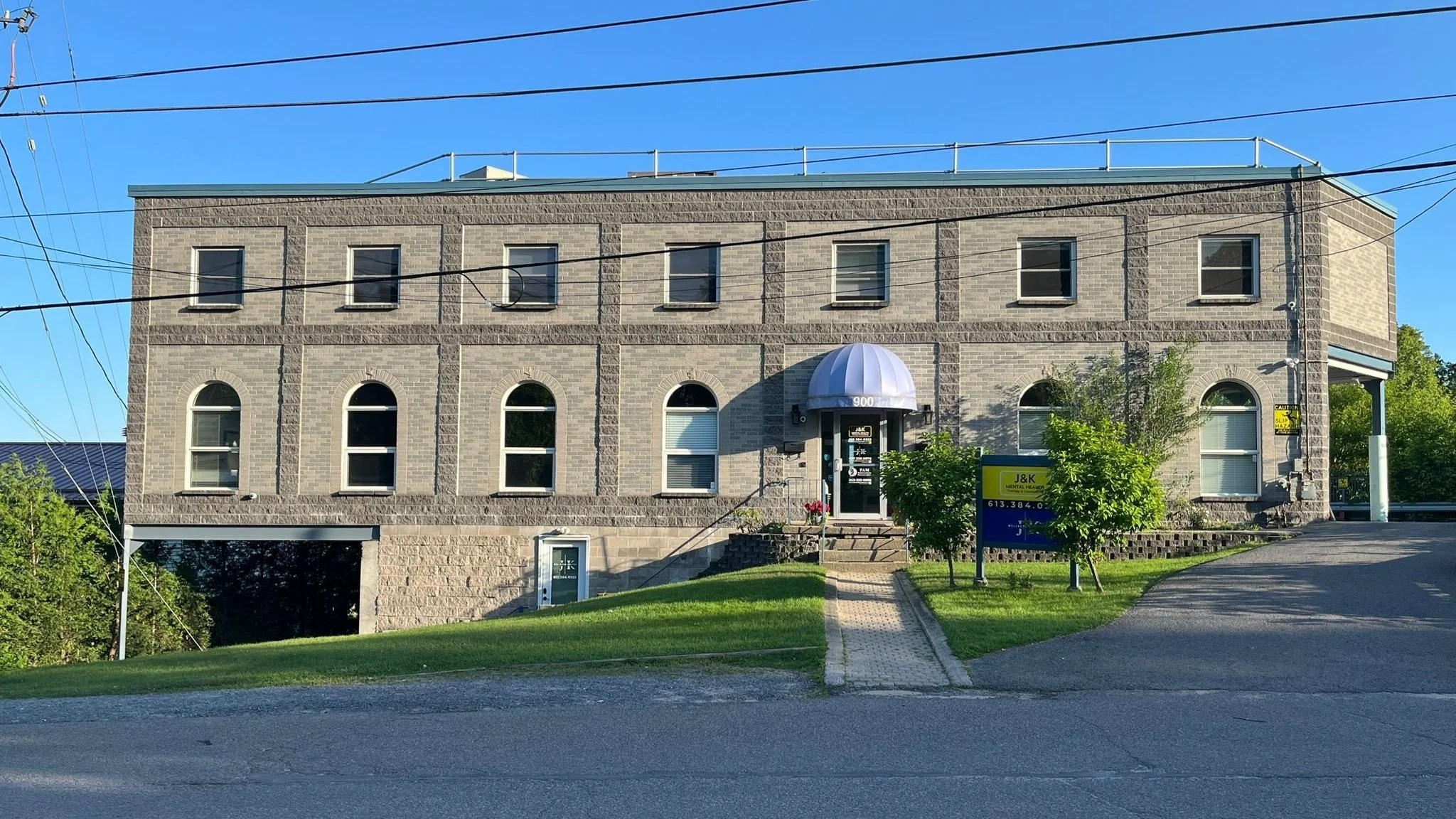 A three-story brick building with arched and rectangular windows, a rounded awning over the entrance, and a sign with contact information in front. There are power lines overhead and a green lawn with trees around.