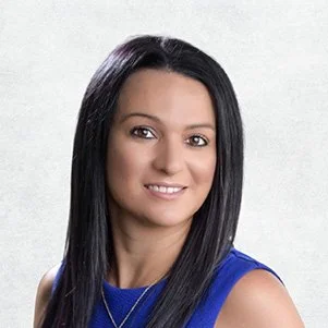 Portrait of a woman with straight black hair, brown eyes, wearing a blue sleeveless top and a gold necklace, smiling against a light background.