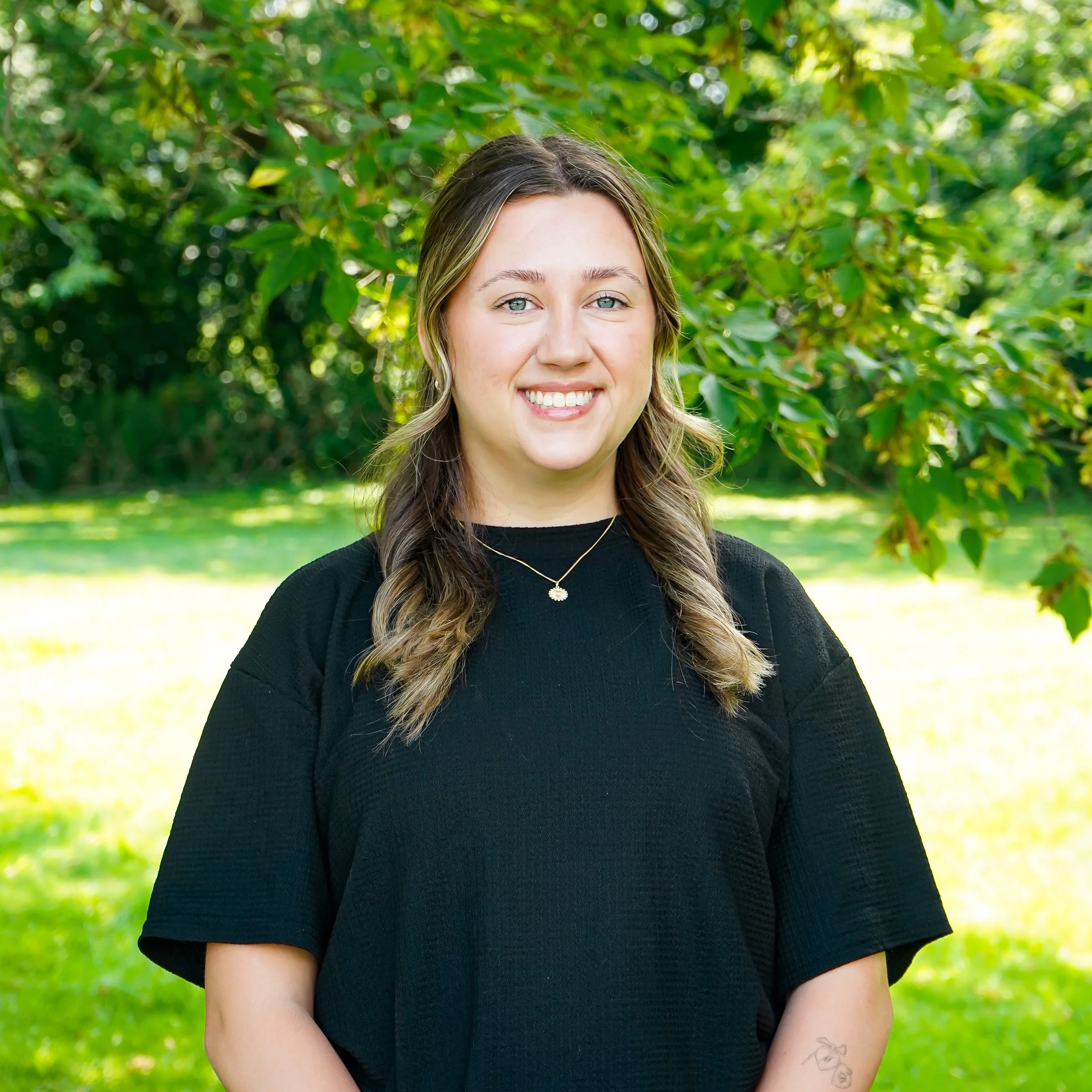 A young woman with long light brown hair, wearing a black top and layered necklaces, smiling at the camera against a plain white background.