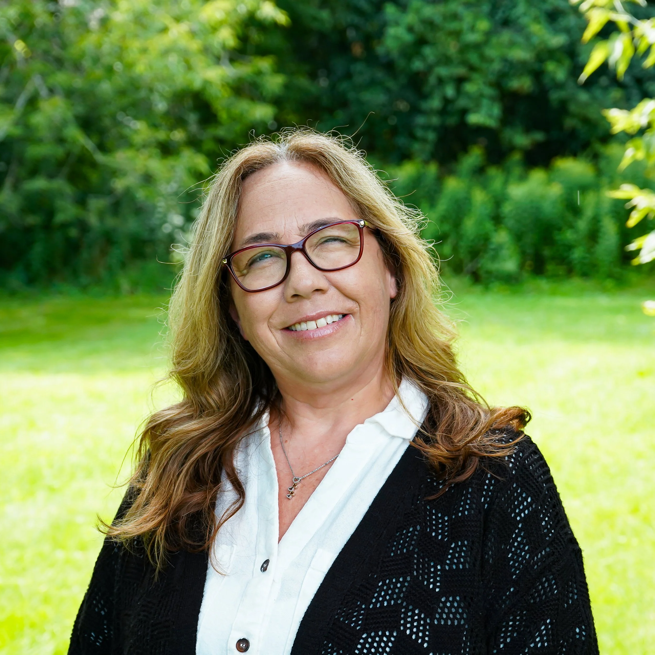 A smiling woman with long brown hair, wearing glasses, a green jacket, and a black top, smiling against a light background.
