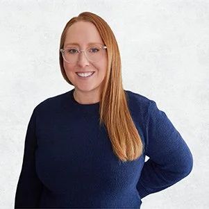 A woman with straight, long red hair and glasses, wearing a navy blue sweater, smiling with her teeth showing, standing against a plain white background.