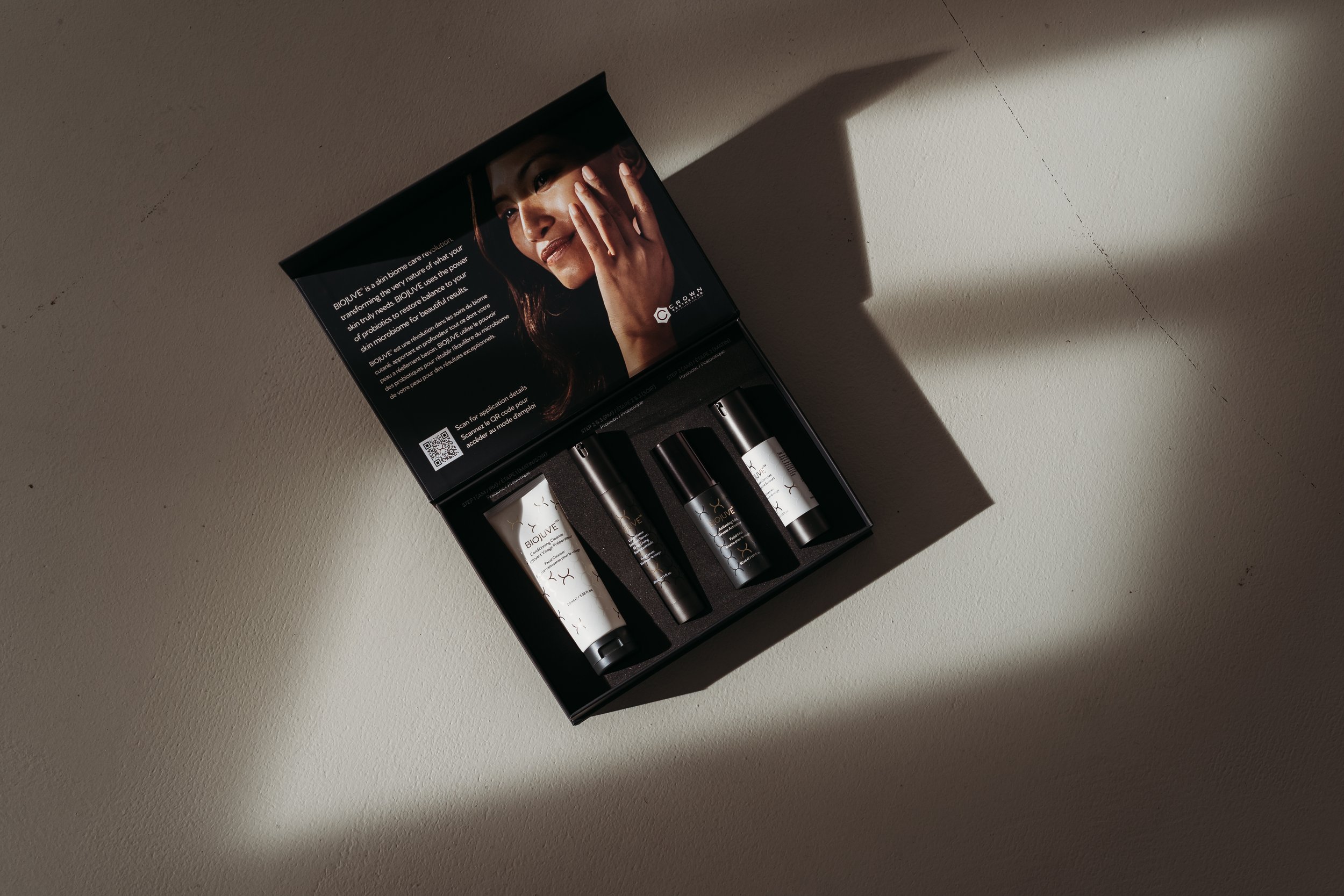 Offener Kosmetikset mit vier Produkte in einer schwarzen Verpackung auf einer hellen Tischfläche, Schatten durch Licht auf der rechten Seite.