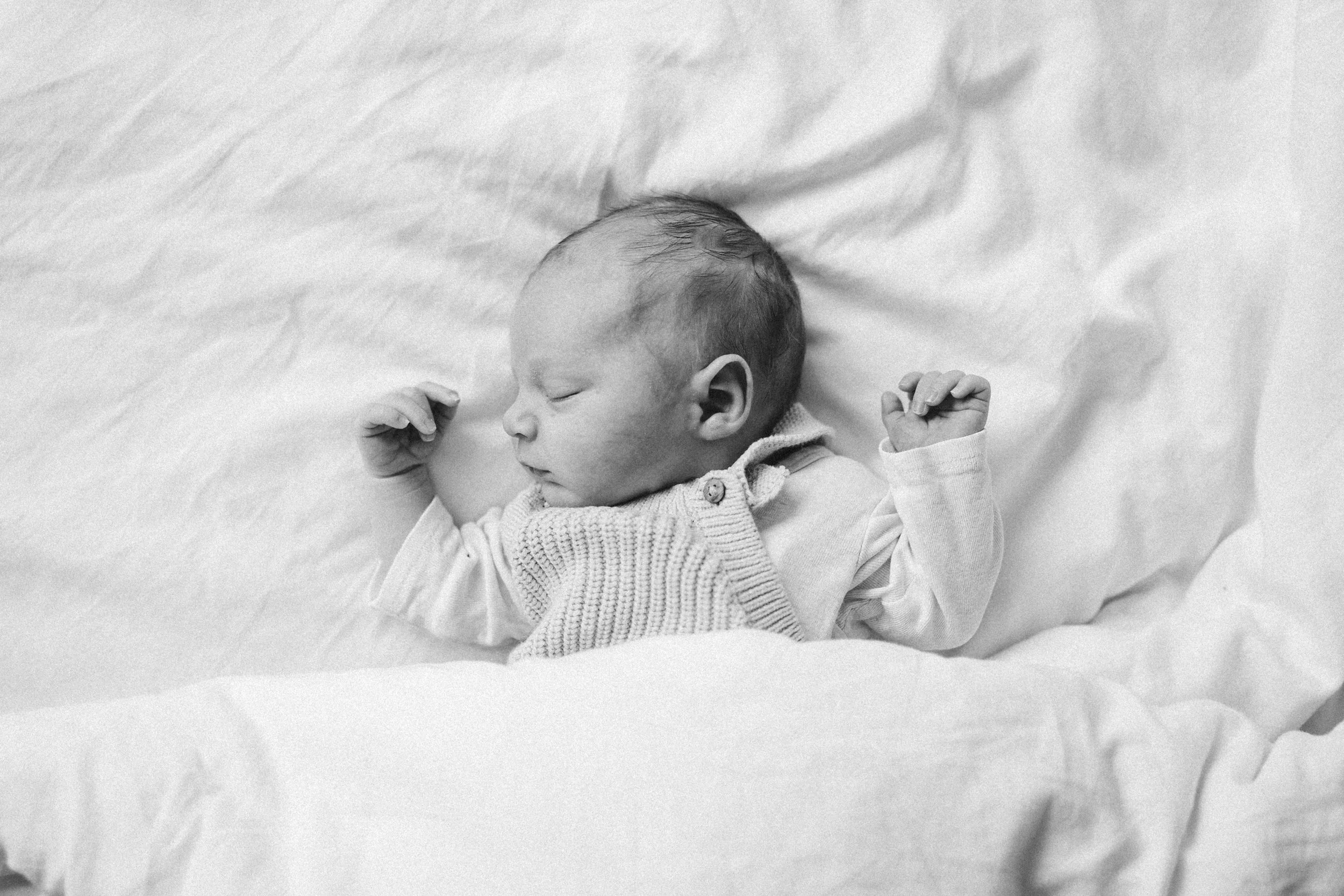 Ein schlafendes Baby, in einem weißen Pullover auf einem weichen Bett mit weißen Bettlaken.