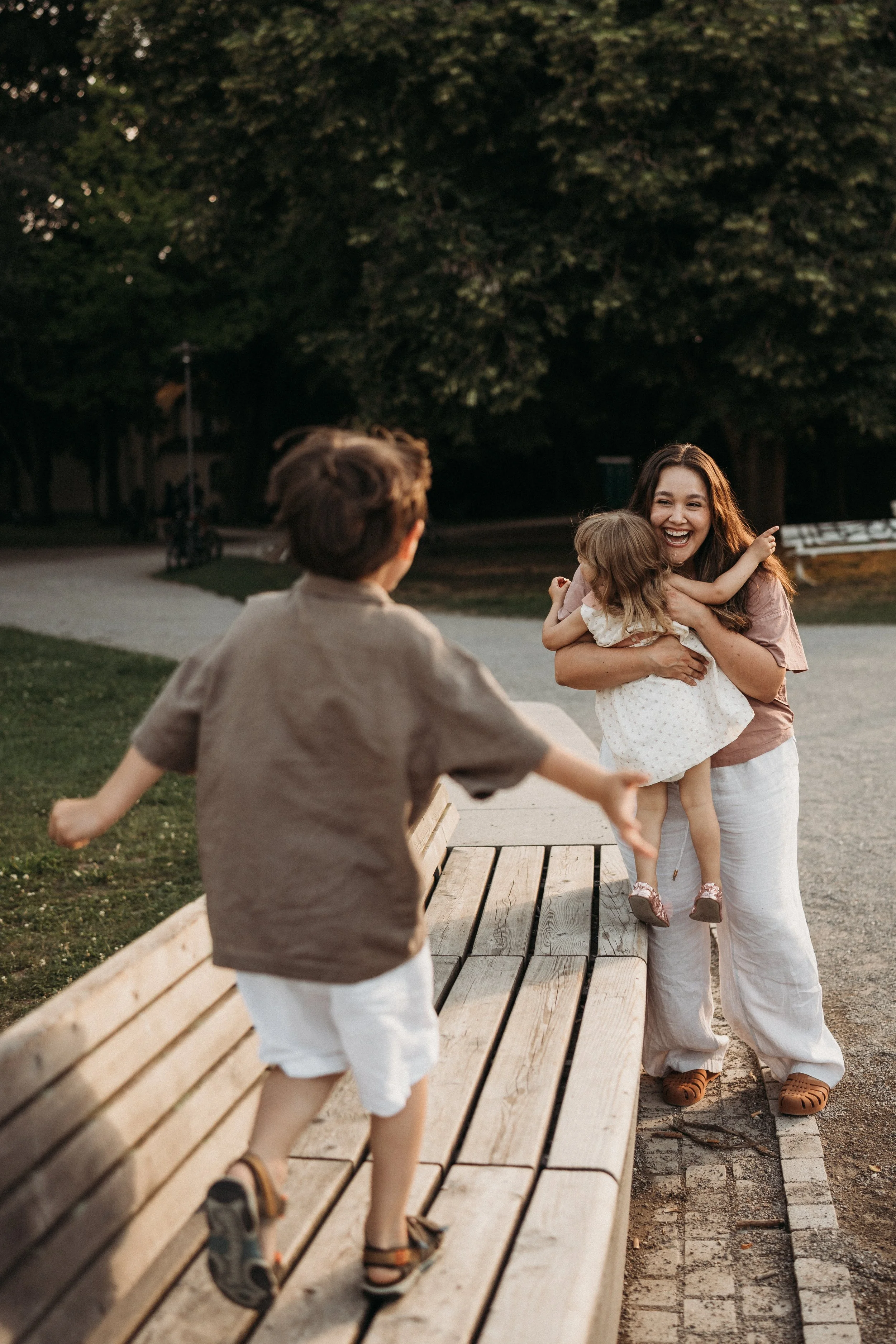Eine Frau und zwei Kinder spielen im Park, die Frau hält ein Mädchen in den Armen, während ein Junge auf einer Bank im Vordergrund läuft.