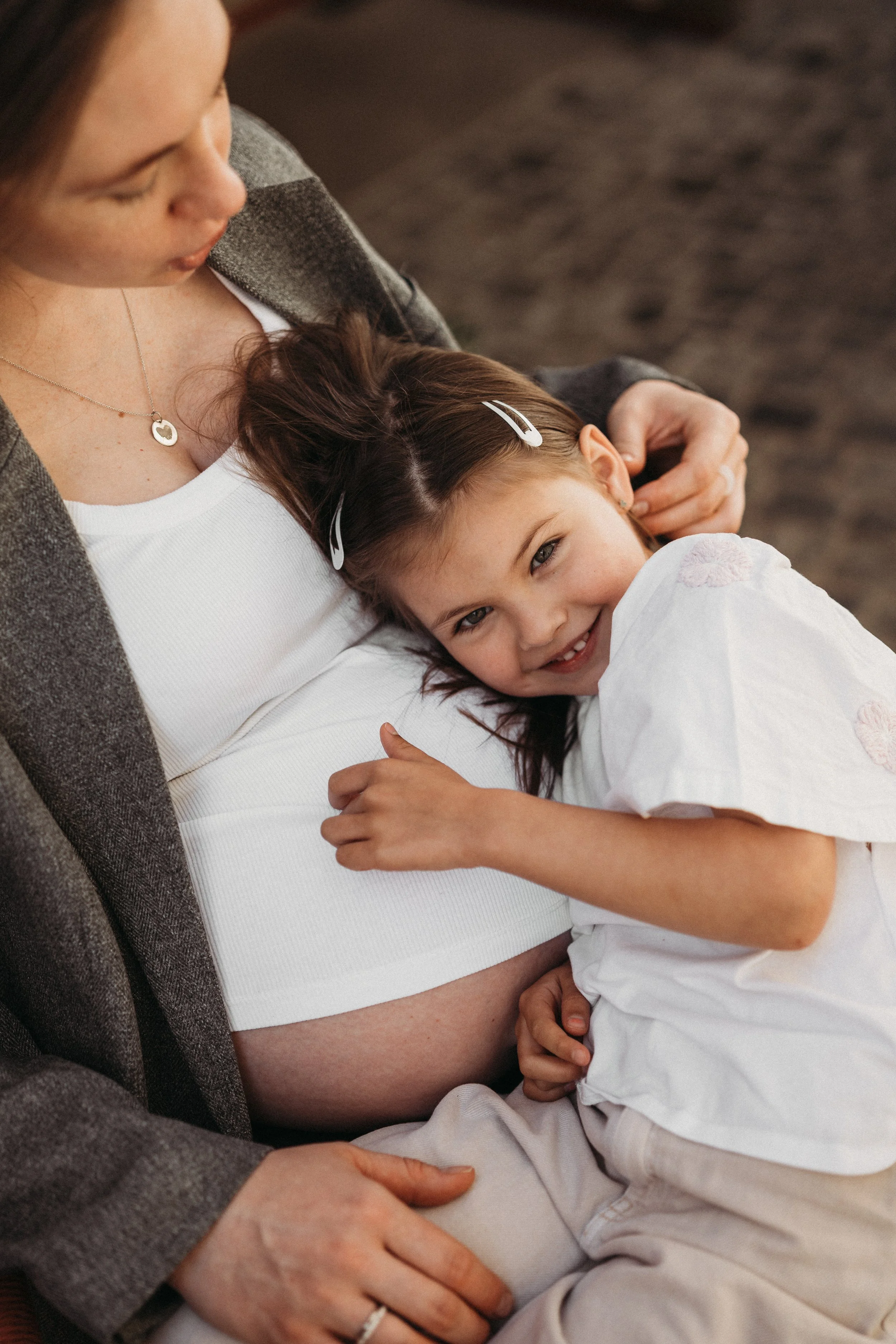 Eine schwangere Frau sitzt auf einem Stuhl, wird von einer jungen Mädchen umarmt und streichelt ihren Bauch. Beide scheinen glücklich und liebevoll miteinander verbunden.