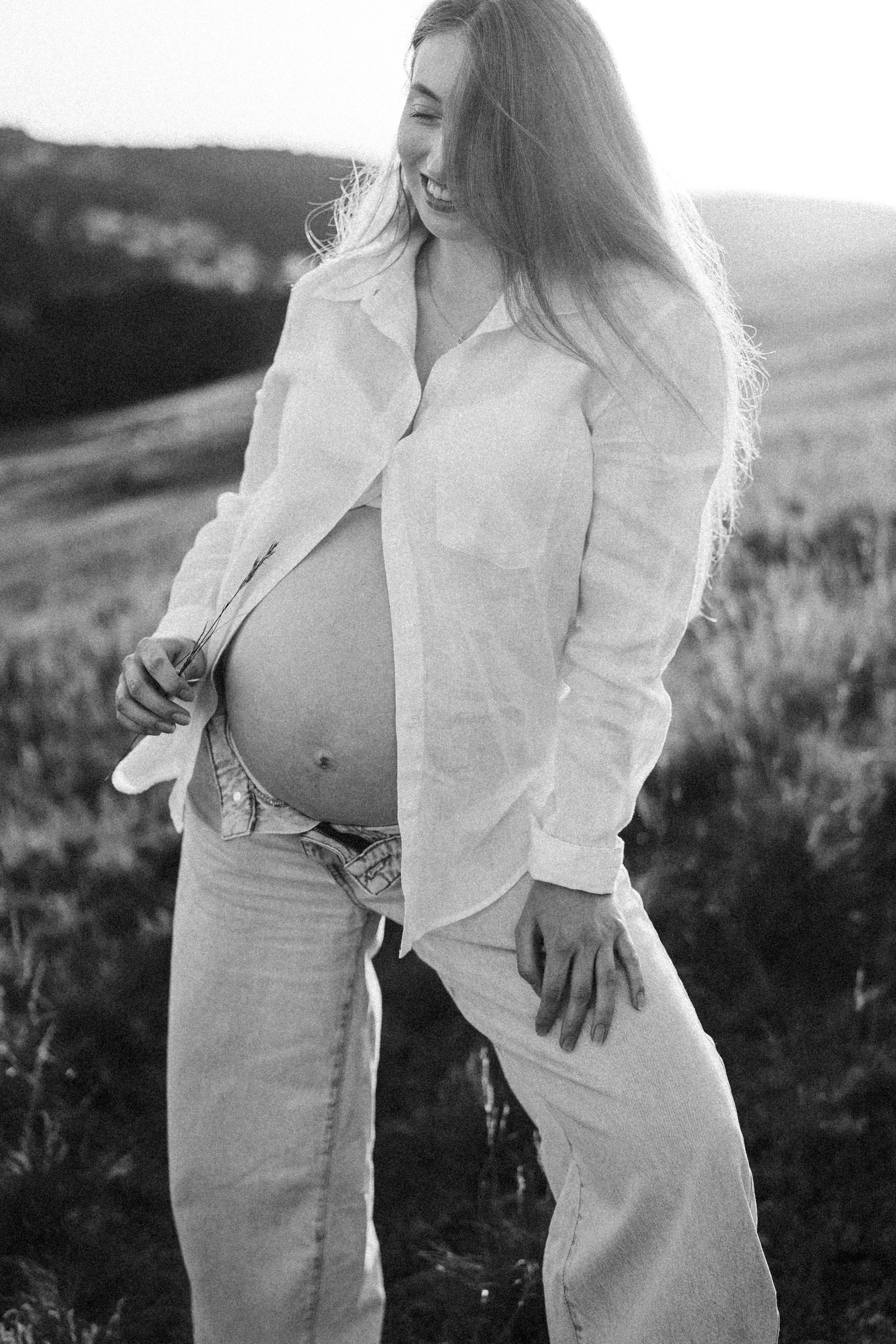 Junge schwangere Frau im Freien, lächelnd, trägt ein offenes Hemd und Jeans, hält eine Blume in der Hand, mit einer ländlichen Landschaft im Hintergrund, in Schwarz-Weiß