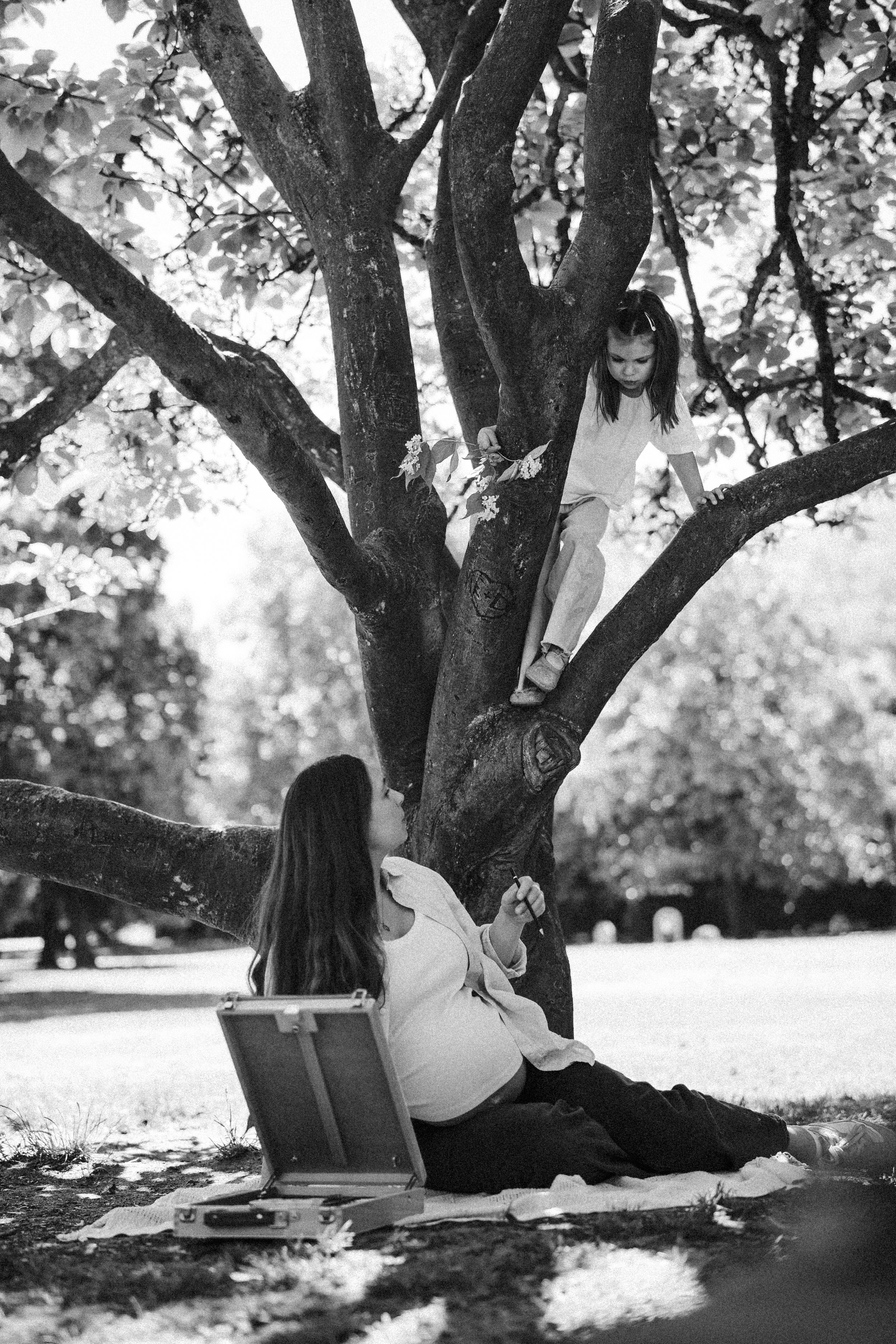 Ein Mädchen klettert in einem Baum, während eine Frau auf dem Boden sitzt und mit einem kurzen Werkzeug arbeitet. Es ist eine parkähnliche Umgebung mit Bäumen im Hintergrund.