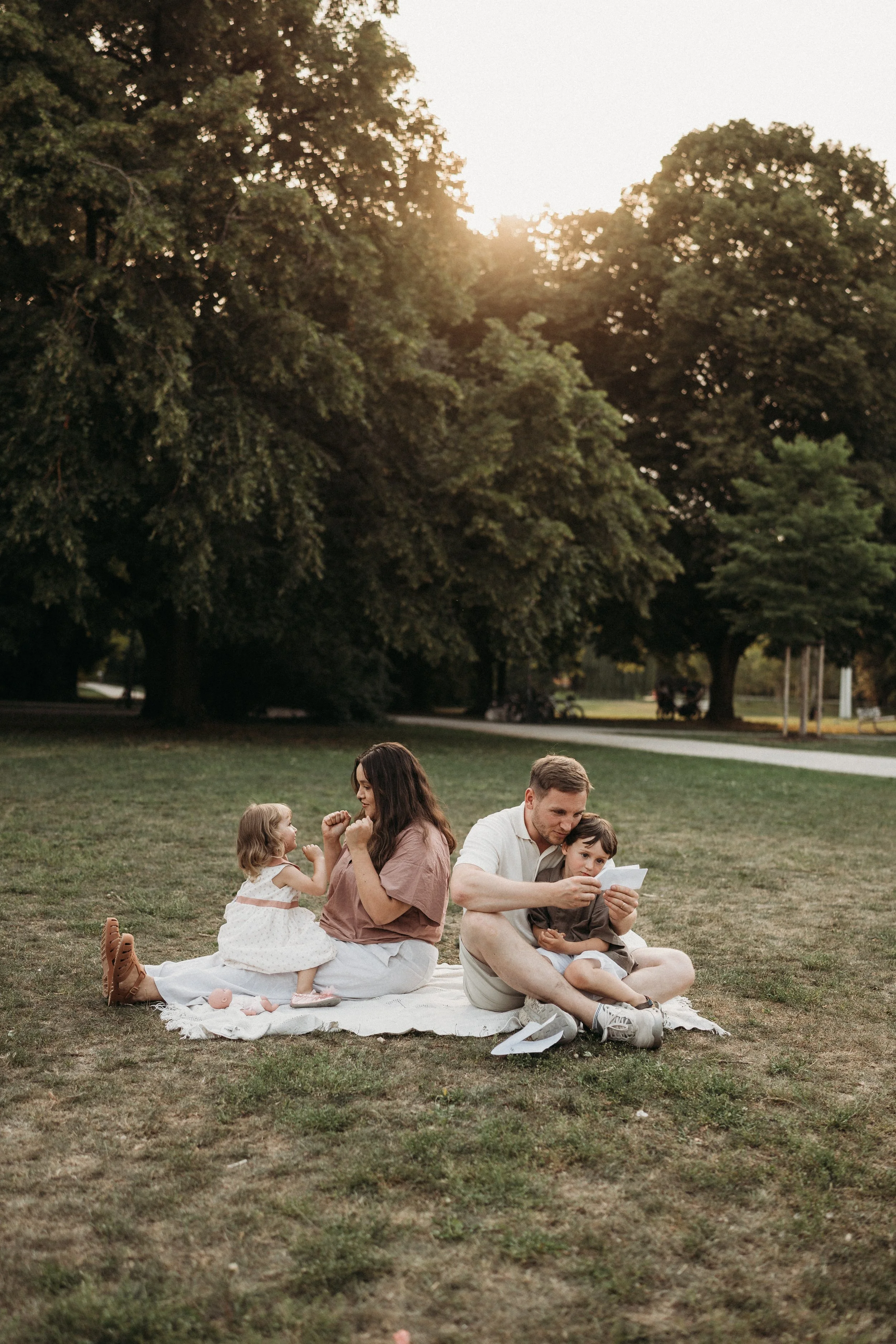 Familie mit zwei Erwachsenen und zwei Kindern sitzt auf einer Decke im Park, liest und spielt, bei Sonnenuntergang, umgeben von Bäumen.