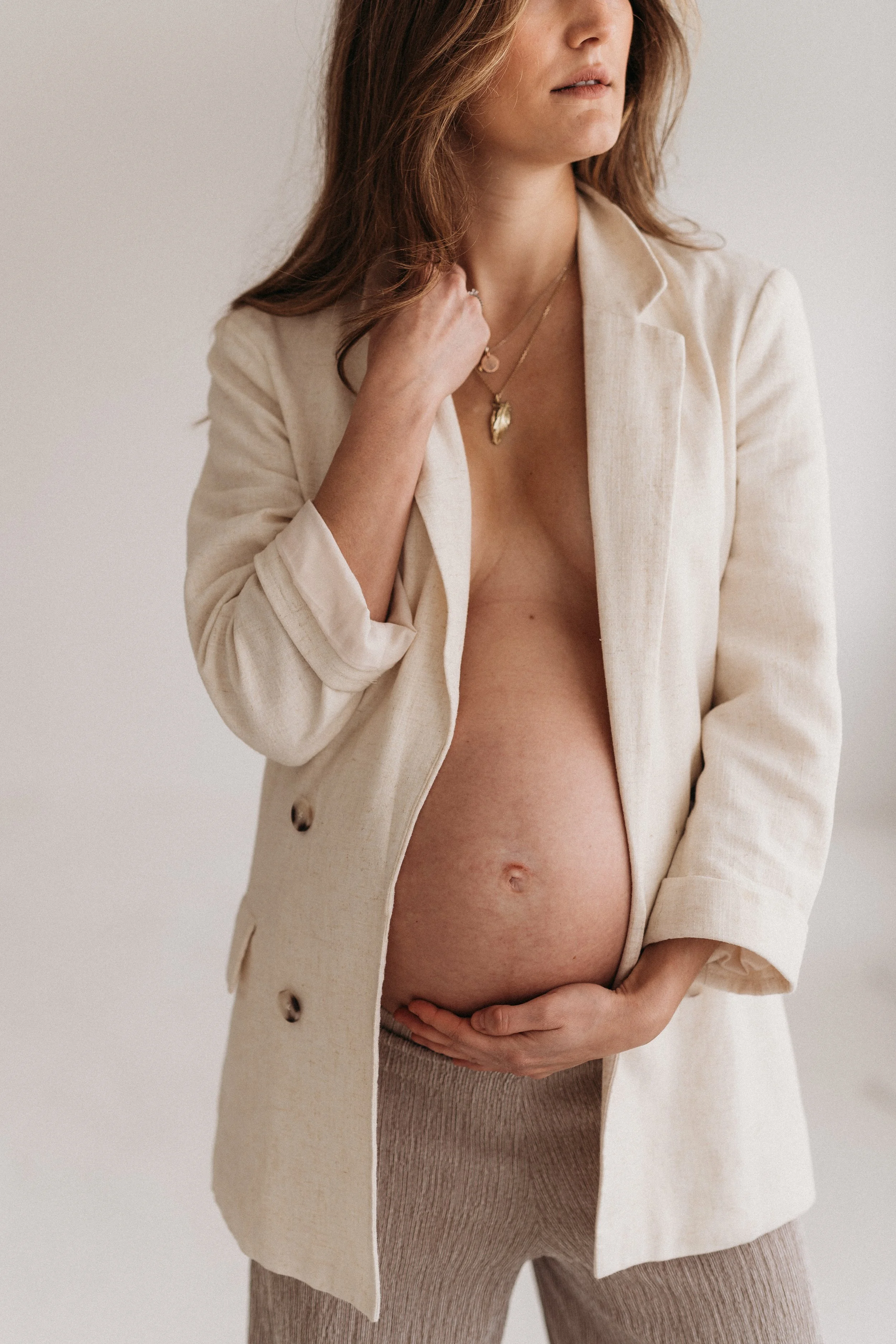 Schwangere Frau trägt beige Jacke und beige Hose, hält Bauch mit einer Hand und berührt ihr Gesicht mit der anderen Hand, trägt Schmuck, Hintergrund ist neutral.