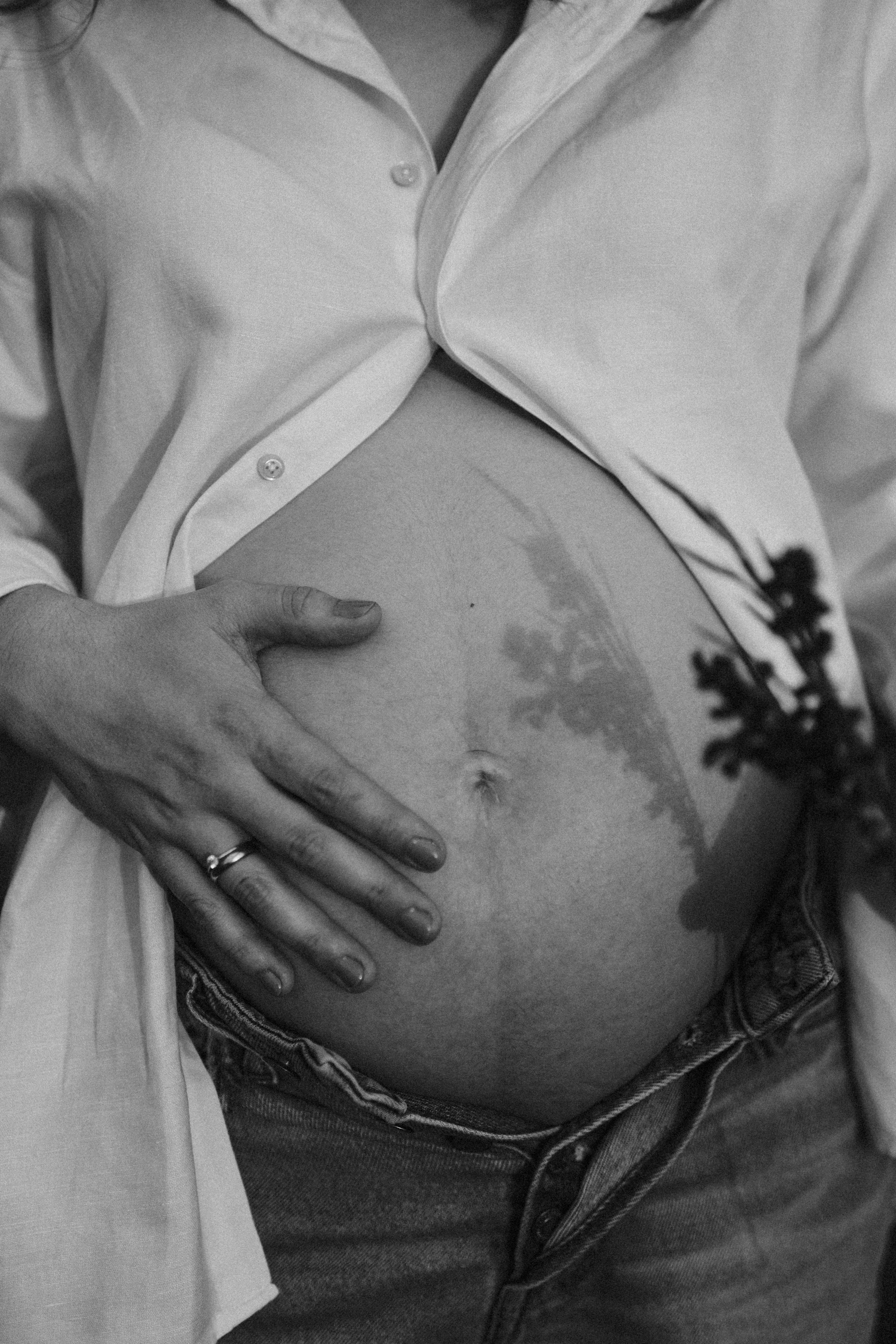 Nahaufnahme eines schwangeren Bauches, eine Hand liegt darauf, eine weitere Hand berührt den Bauch. Es ist schwarz-weiß fotografiert.