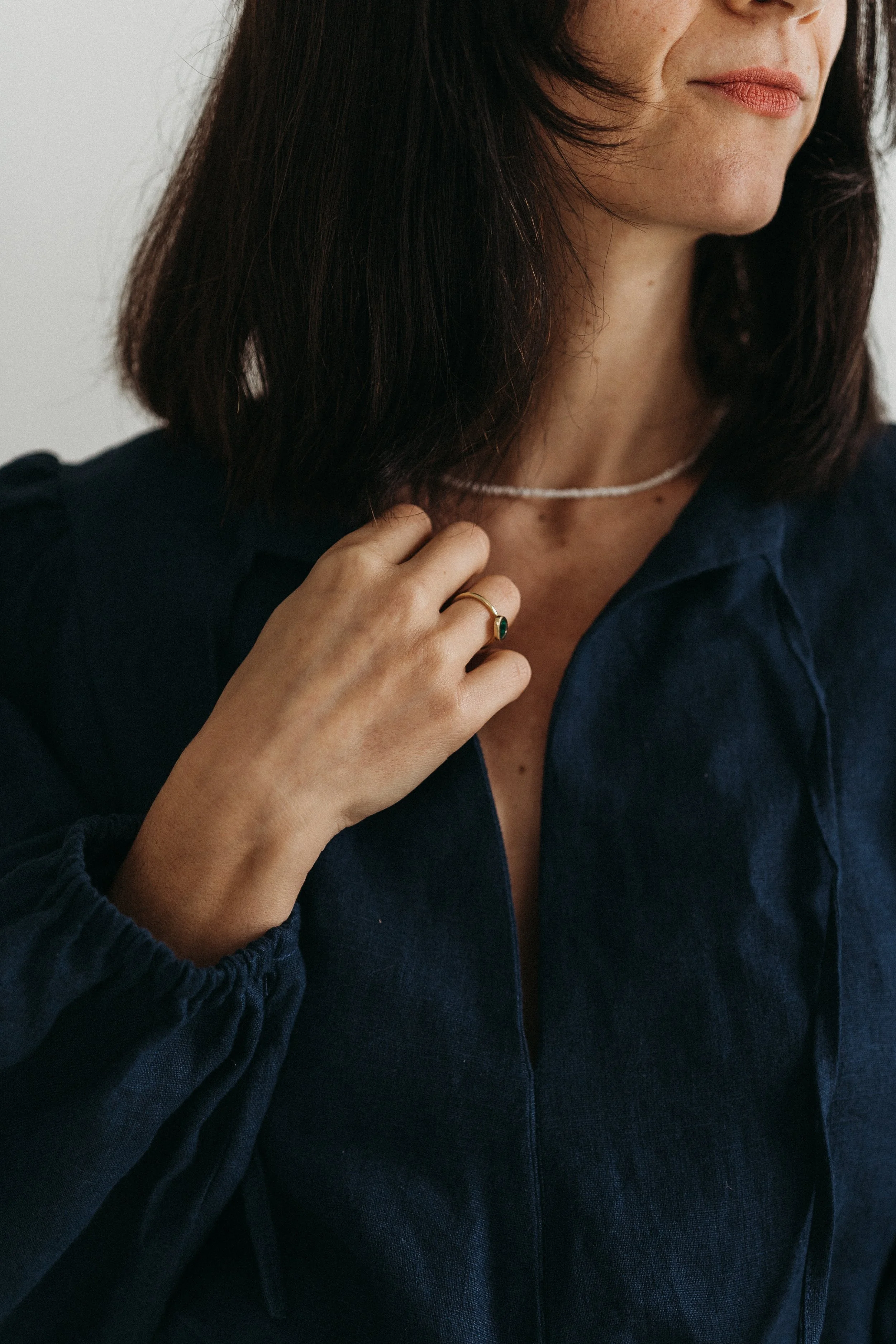 Nahaufnahme einer Frau, die eine Hand an die Schulter legt, trägt einen goldenen Ring mit einem grünen Edelstein und eine Perlenkette, trägt eine dunkelblaue Bluse, ausgeglichener Gesichtsausdruck.