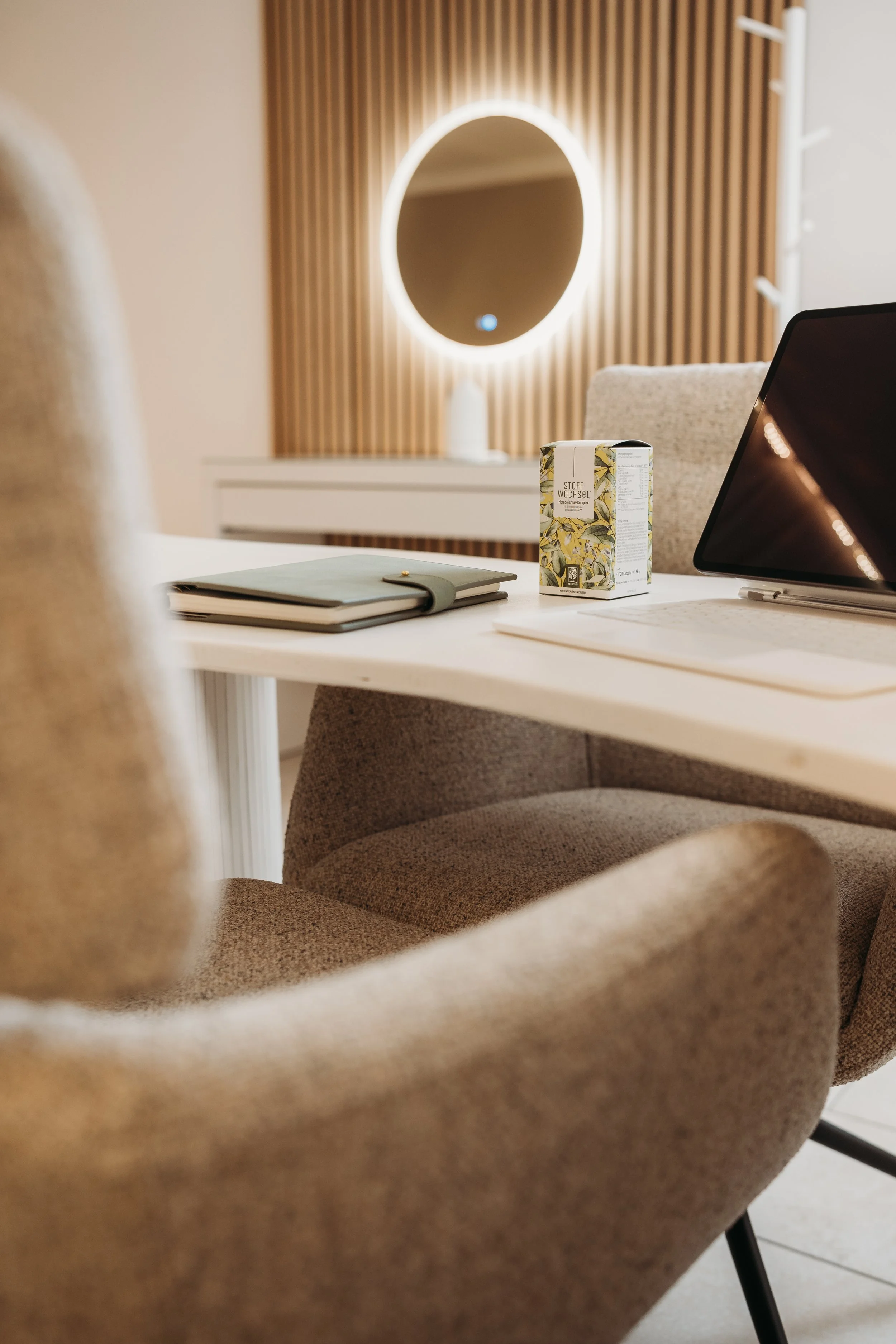 Arbeitsplatz mit Stift, Notizbuch, Kiste mit Tee, Laptop und Notizblock auf einem weißen Tisch. Im Hintergrund ein runder Spiegel mit Licht, an einer Wand mit Holzvertäfelung.