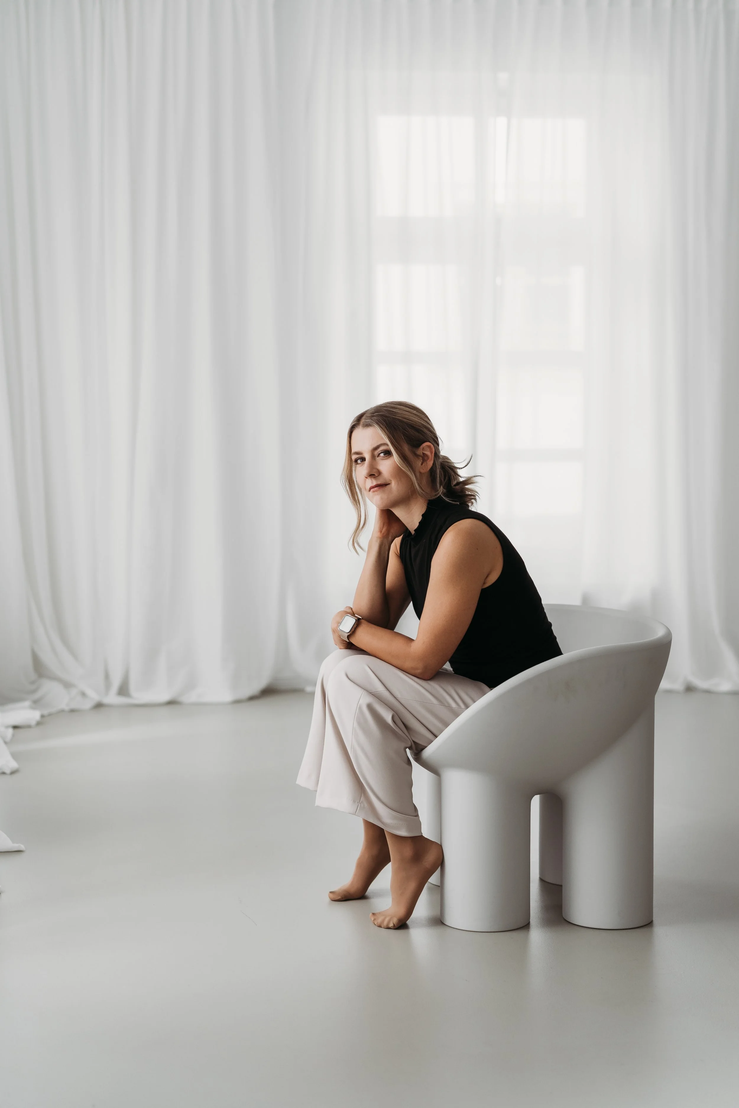 Eine Frau sitzt auf einem modernen weißen Stuhl in einem hellen Raum mit weißen Vorhängen im Hintergrund.