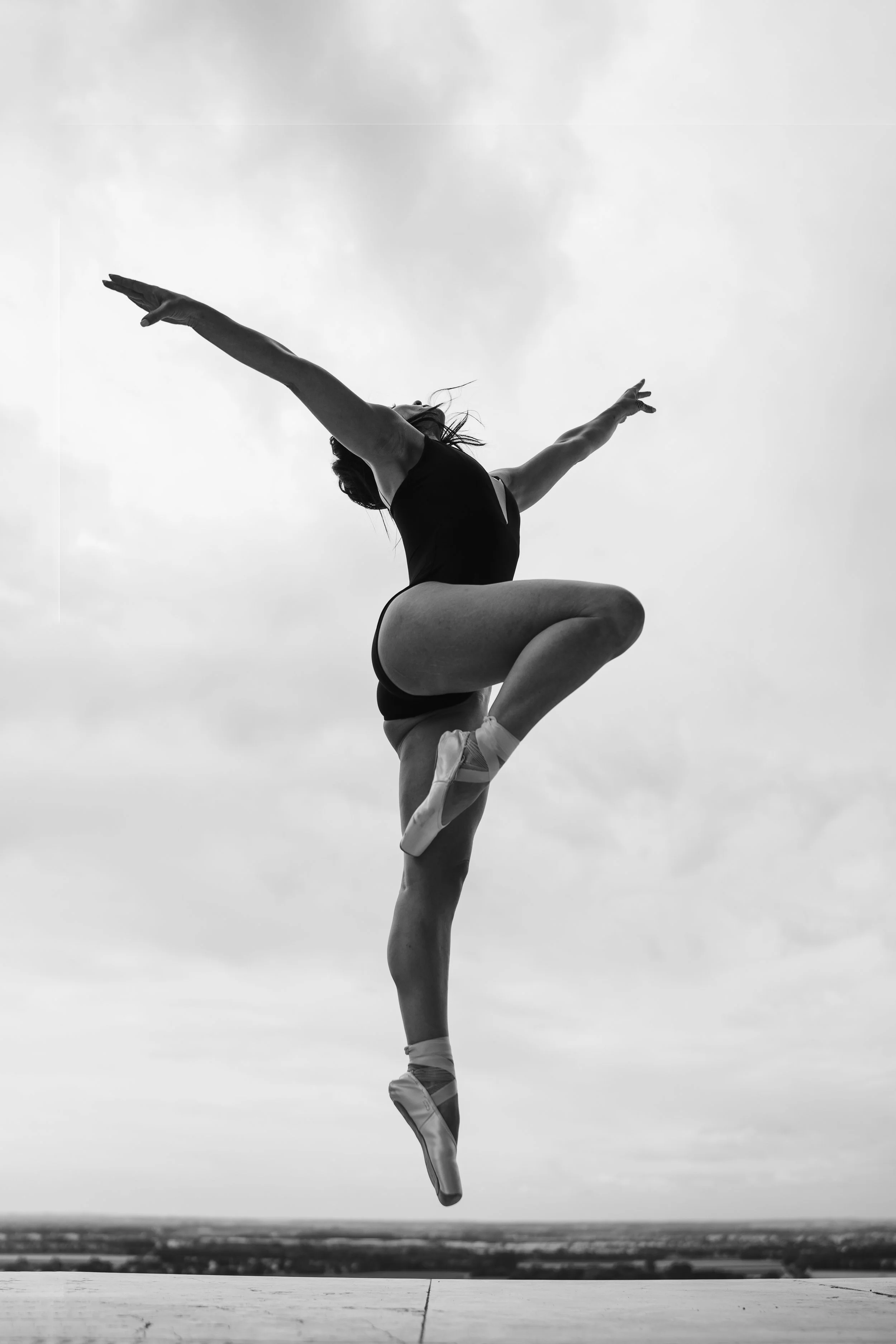 Schwarzweißfoto eines Balletttänzers, der in der Luft posiert, mit einem Bein auf dem Boden und dem anderen Bein angewinkelt. Die Arme sind ausgestreckt und der Himmel im Hintergrund ist bewölkt.